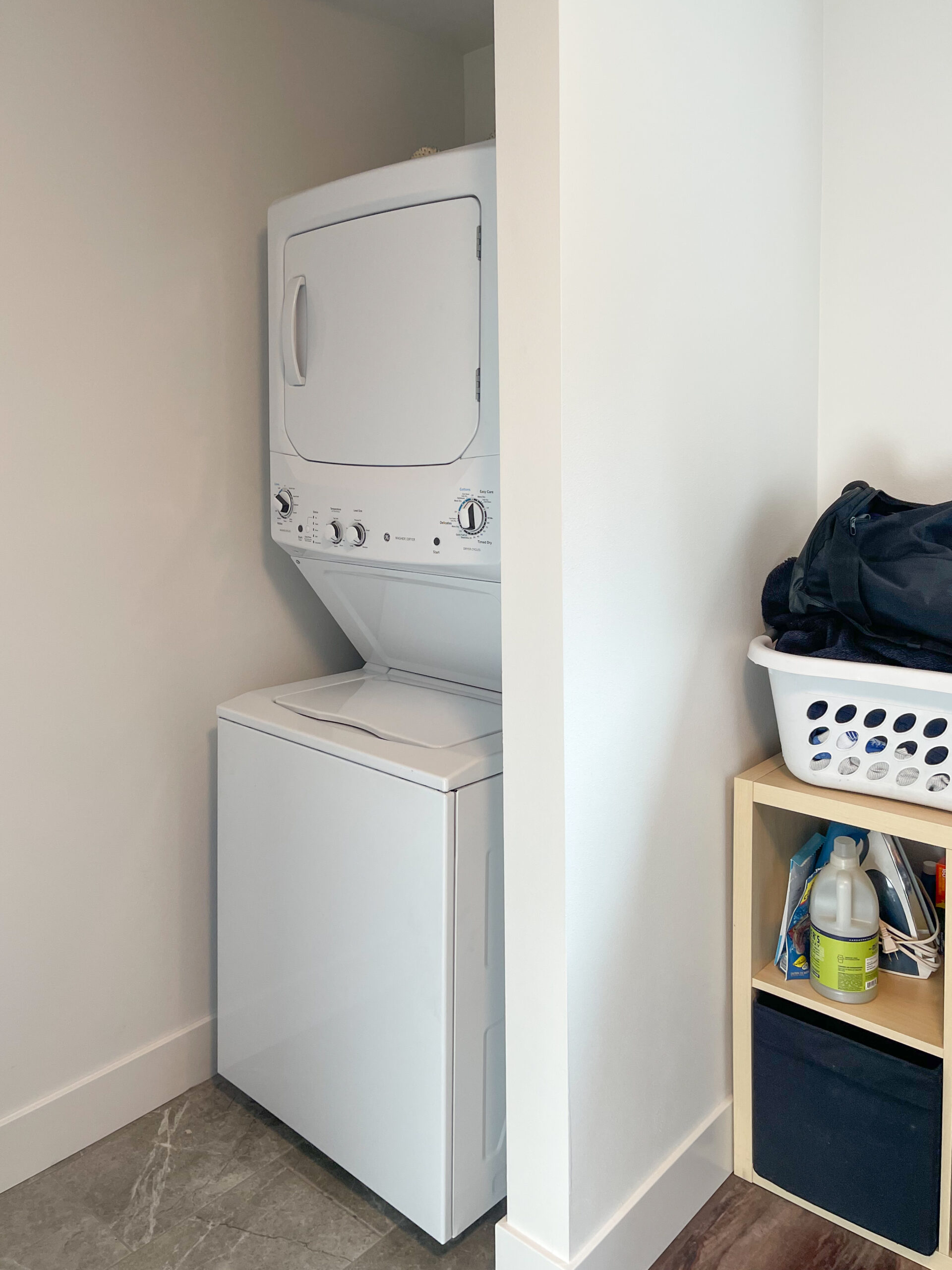 A compact laundry area with a stacked white washer and dryer next to a shelf holding a laundry basket, black bag, and cleaning supplies against neutral-colored walls and tile flooring.