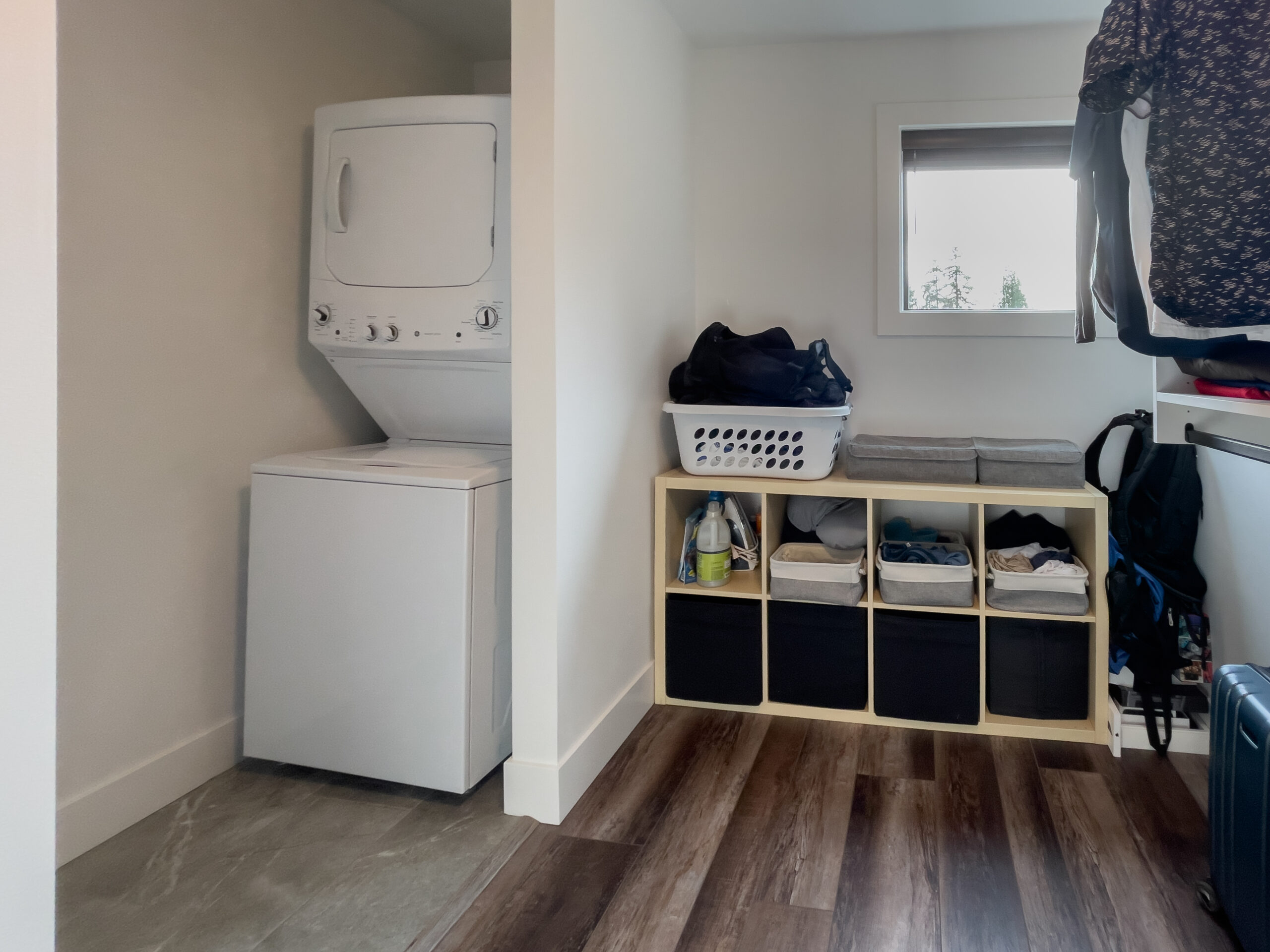A laundry room with a stacked washer and dryer, shelving with cubbies, baskets, folded clothes, and cleaning supplies, and a window letting in natural light.