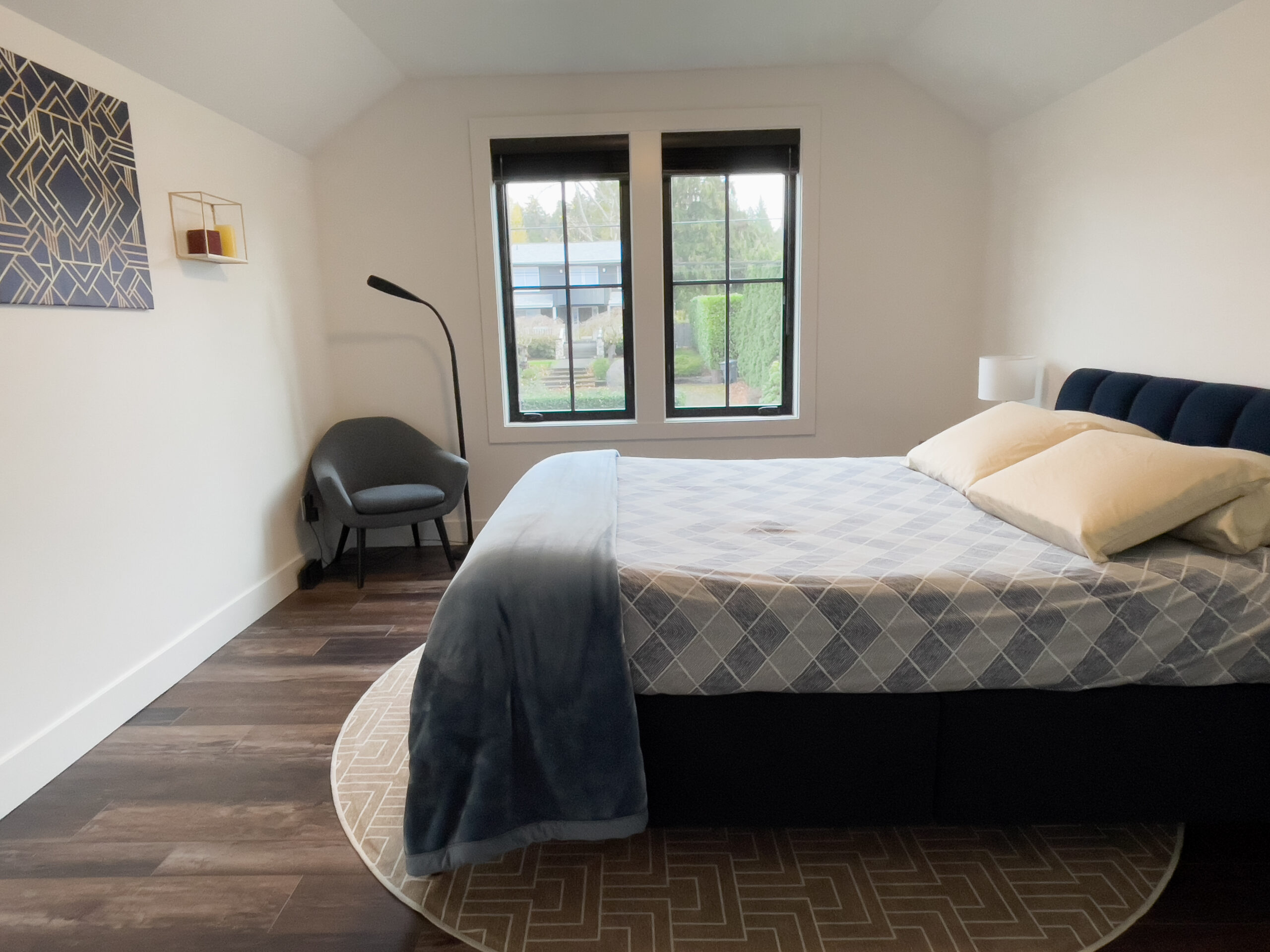 A modern bedroom with a double bed, blue blanket, two pillows, a floor lamp, a gray chair in the corner, and two large windows letting in natural light. The floor is dark wood, and there is minimalistic decor on the walls.