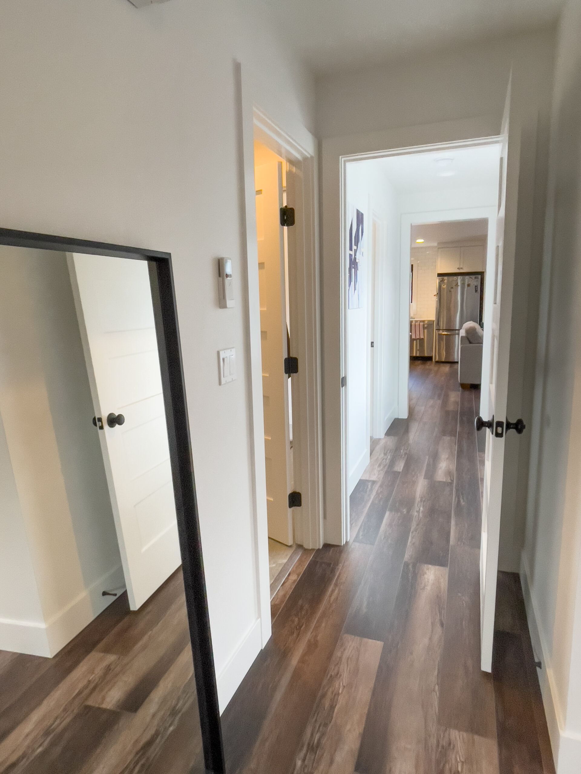 A hallway with dark wood floors, white walls, and white doors. An open door on the left reveals a bathroom, while another open door leads to a kitchen area visible in the background. A mirror stands on the left.