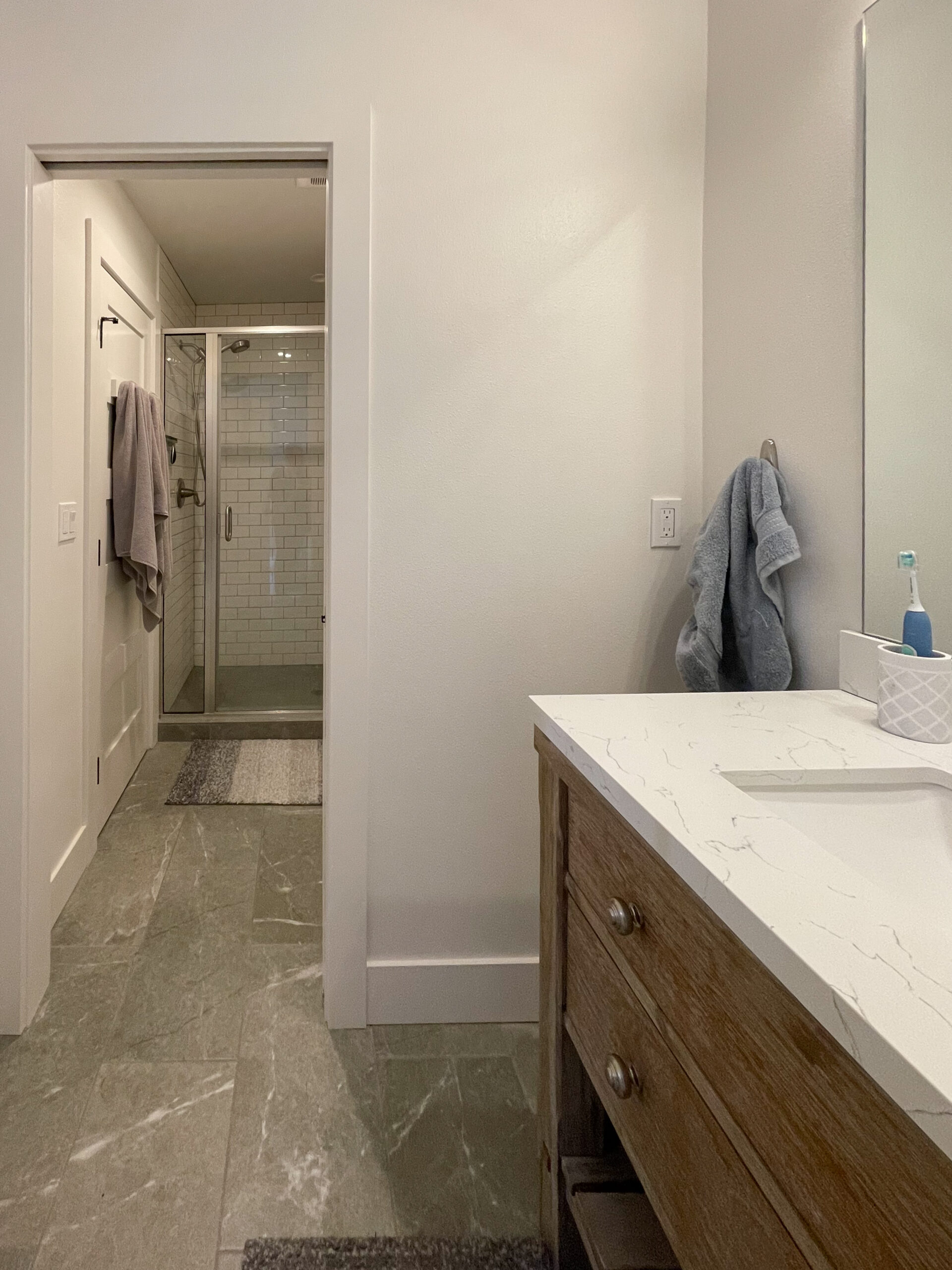 Modern bathroom with gray tile floor, glass-enclosed shower, white vanity countertop, wooden cabinet, large mirror, gray towel on a hook, and toiletries on the sink. Neutral colors and clean, minimal design.