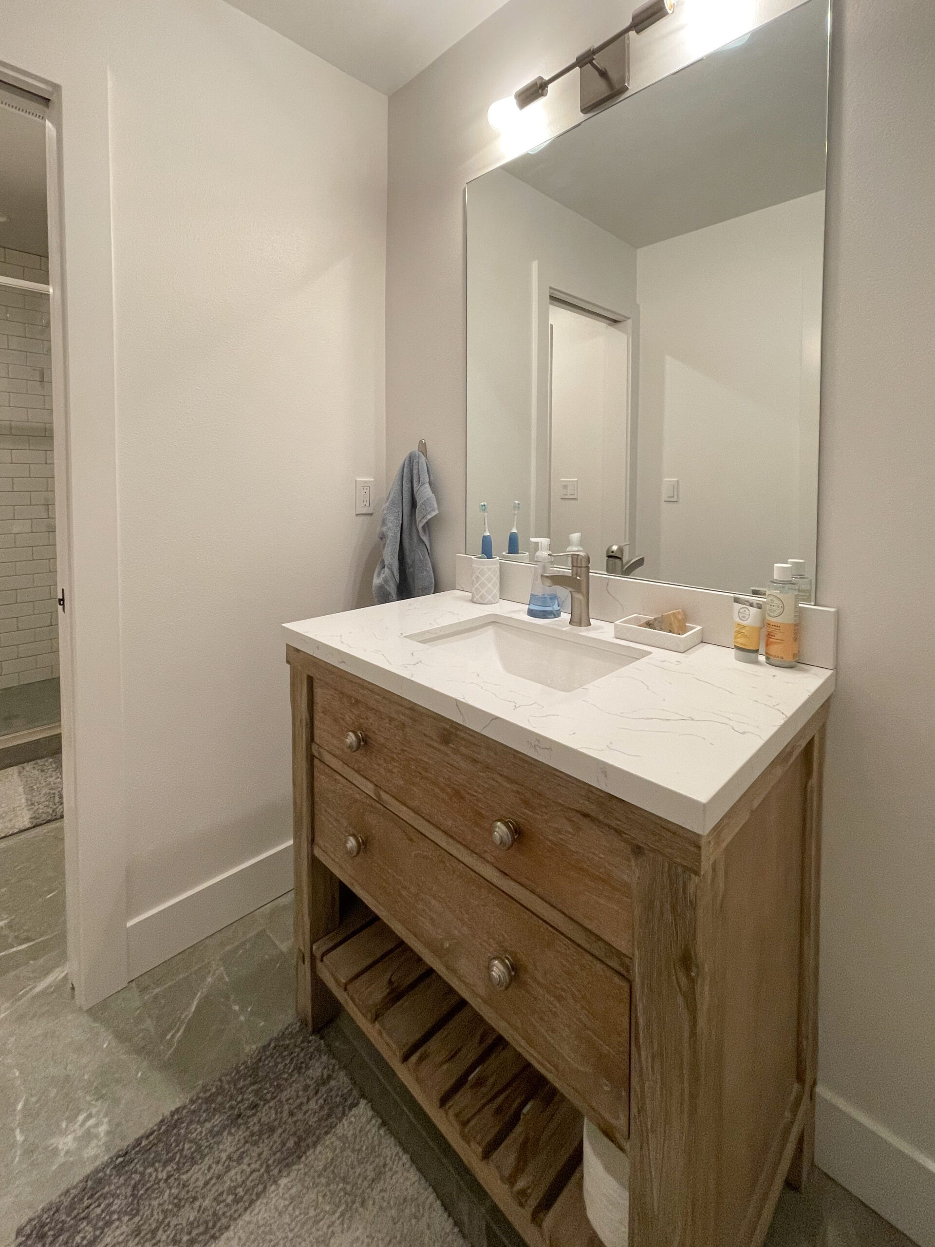 A wooden bathroom vanity with a white countertop, soap dispensers, and toiletries sits below a large mirror. A towel hangs on the wall. The room has white walls and a gray tiled floor.