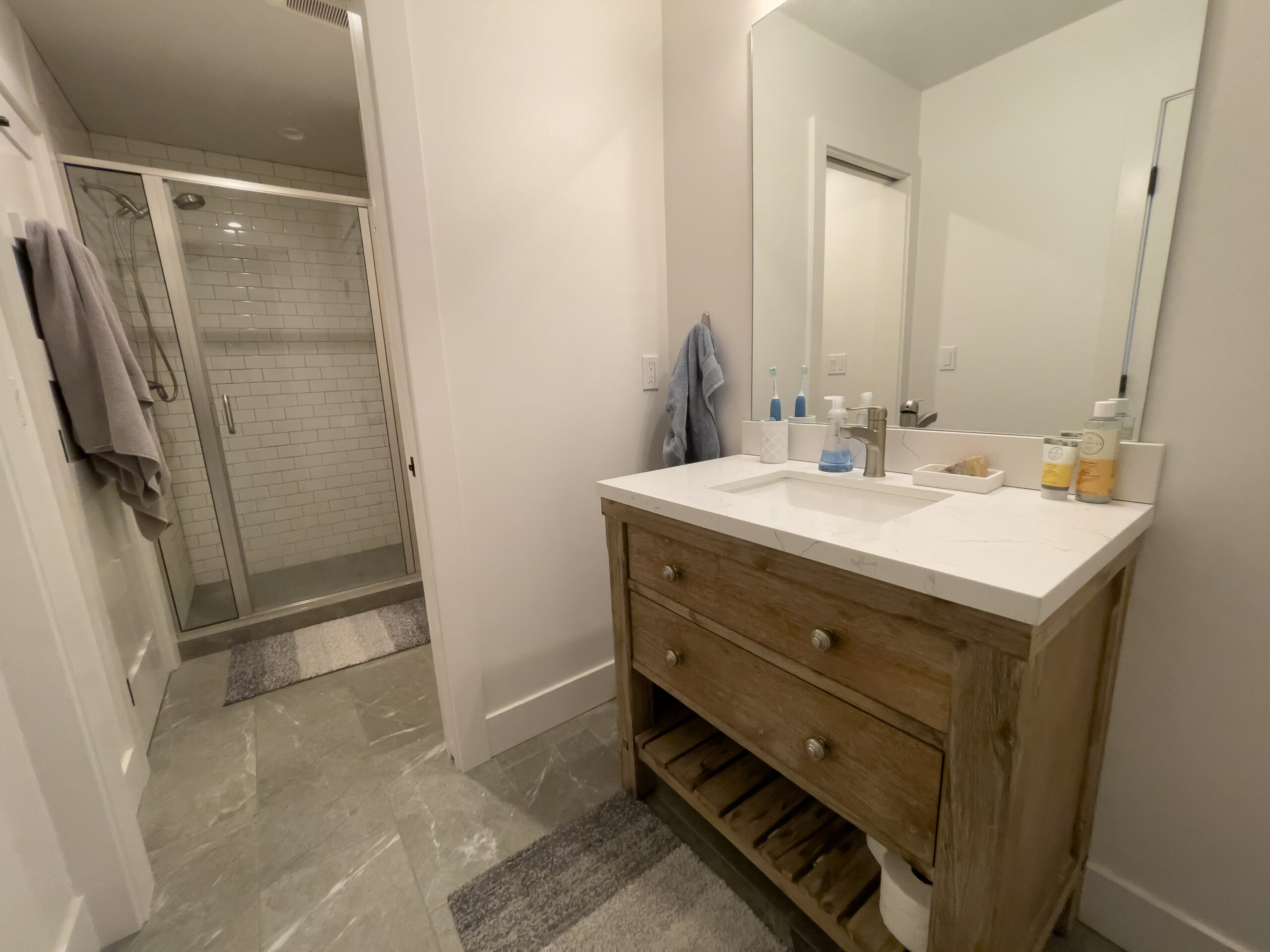 A modern bathroom with a wooden vanity, white countertop, large mirror, and toiletries. In the background, there is a glass-enclosed shower with white subway tiles and two rugs on the gray tiled floor.