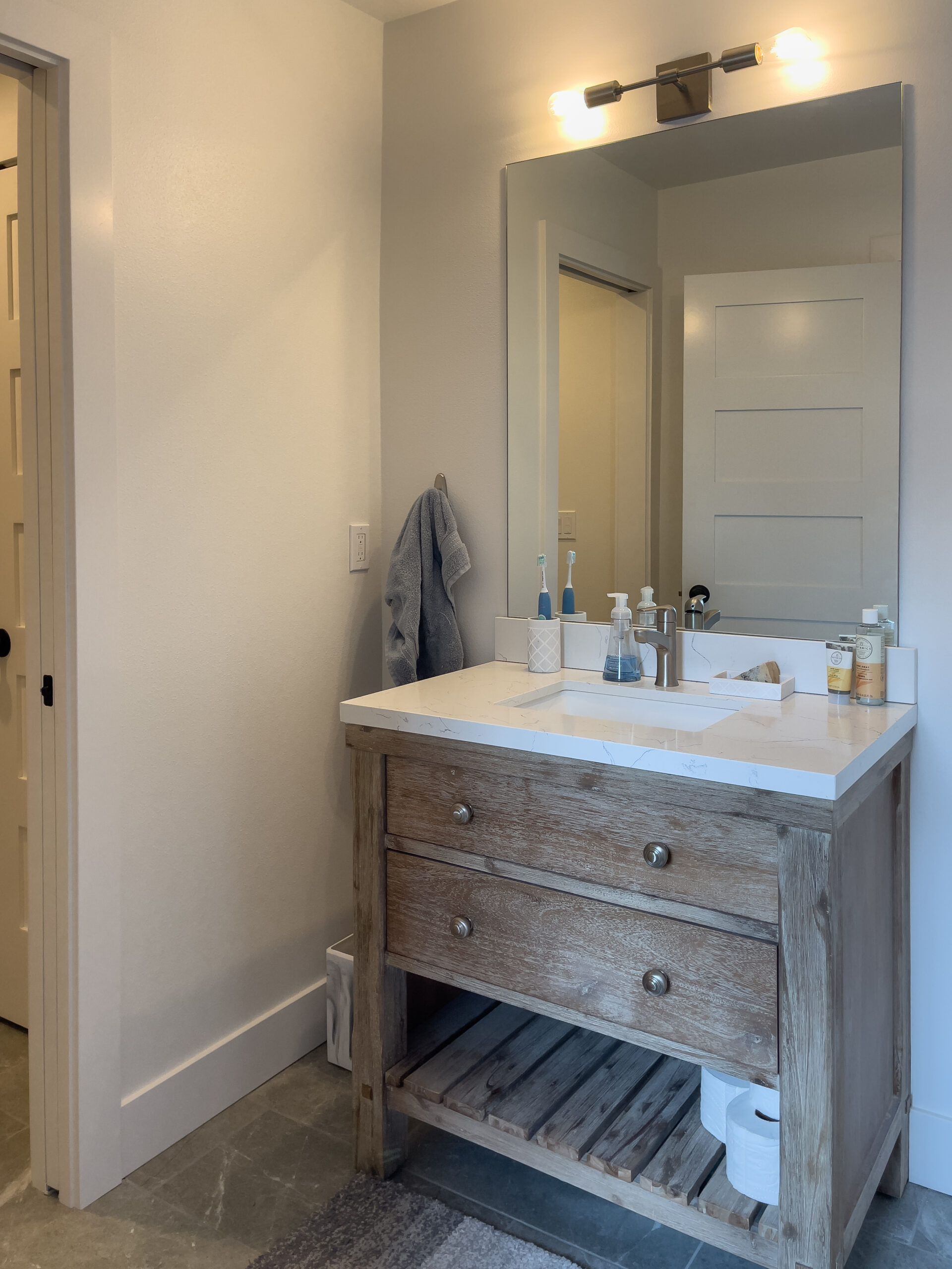 A bathroom with a wooden vanity, white countertop, large mirror, and modern light fixture. Toiletries, soap, and toothbrushes are on the counter. A gray towel hangs on the wall, and a roll of toilet paper is on the lower shelf.