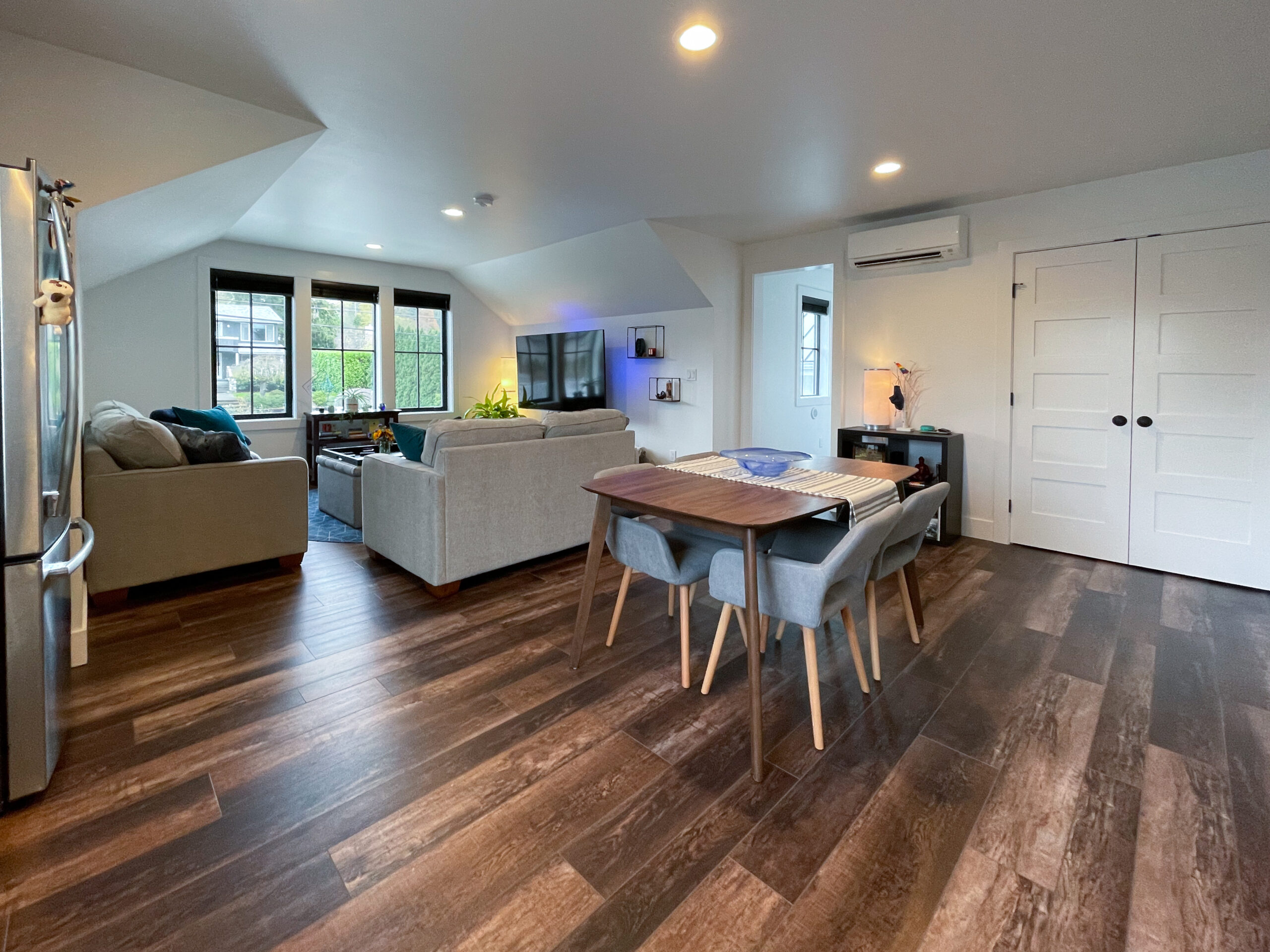 Modern open-plan living and dining area with wooden floors, two beige sofas, a dining table with four chairs, large windows, wall-mounted TV, and white double doors.