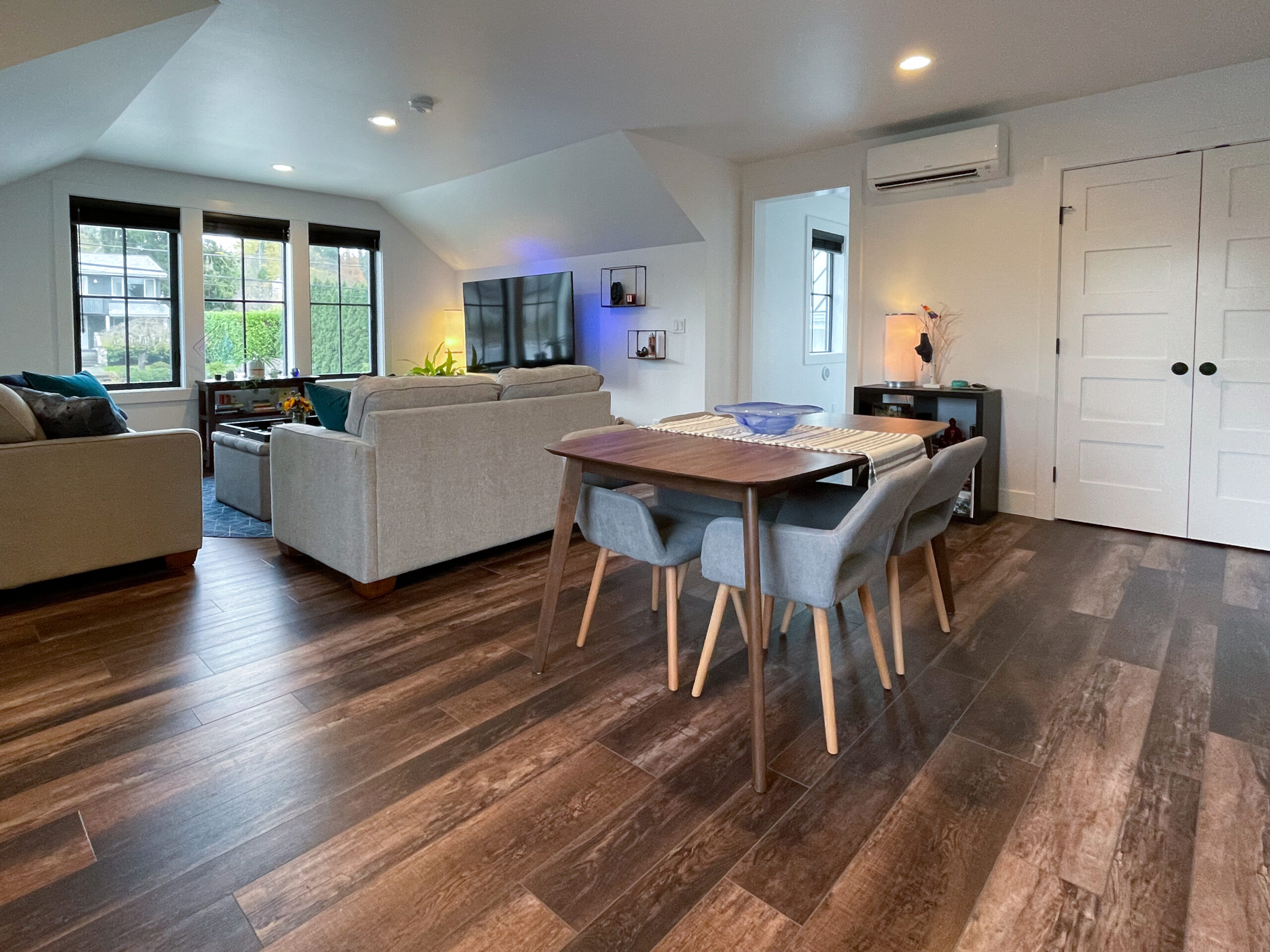 A modern living and dining area with wood floors, a dining table with four chairs, two light gray sofas, a TV on the wall, large windows, and indoor plants. The room is bright and neatly organized.