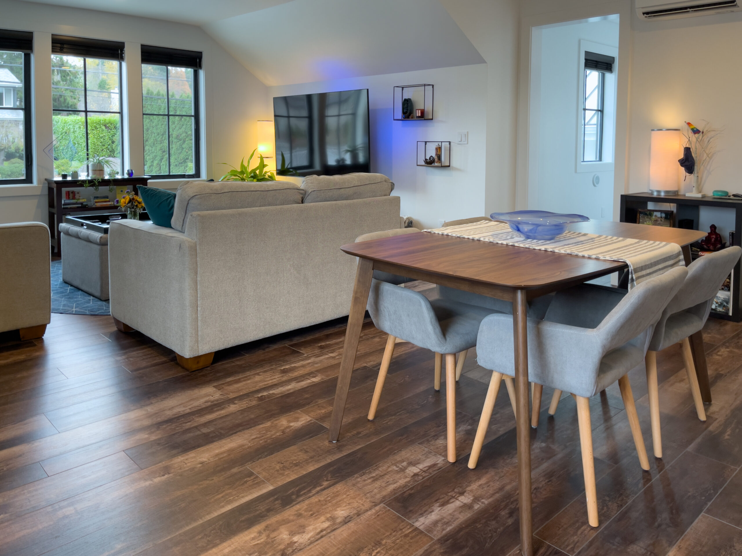 Modern living and dining area with wood flooring, gray sofas, TV on the wall, a dining table with four gray chairs, large windows, and contemporary decor. Natural light fills the room.