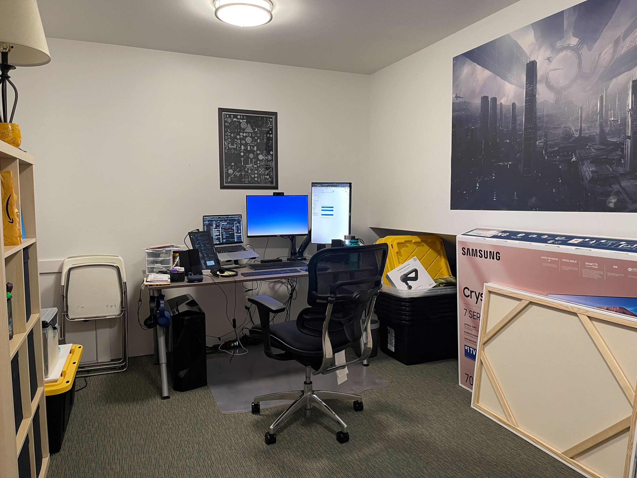 A home office with a desk holding computers, a laptop, monitors, and office supplies. A black chair is at the desk. The room has shelves, storage bins, wall art, and a ceiling light. The space appears organized yet busy.