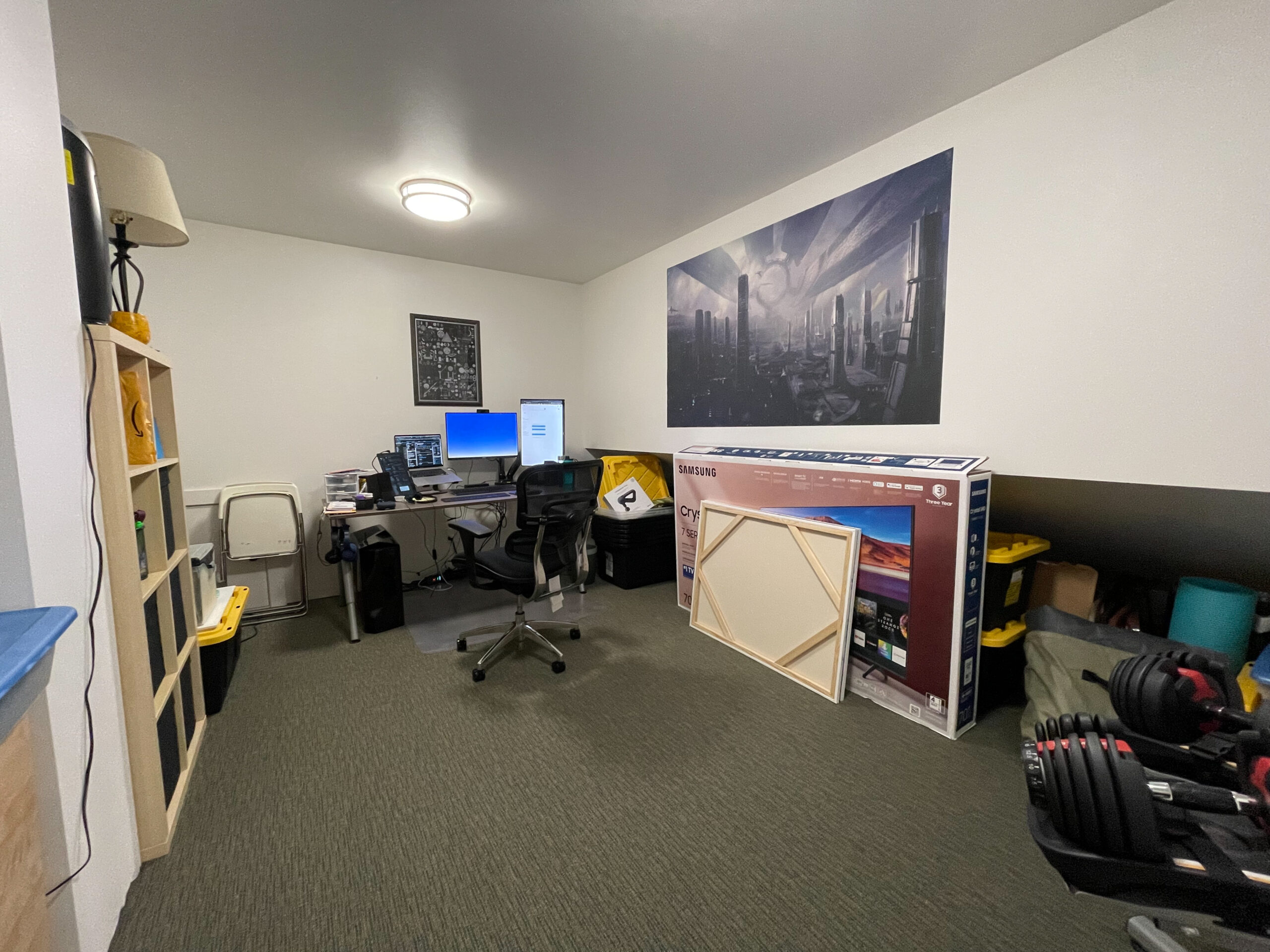 A home office with a desk, two monitors, an office chair, and shelves. A large TV in its box and a framed picture lean against the wall. Dumbbells and storage bins are also visible in the room.