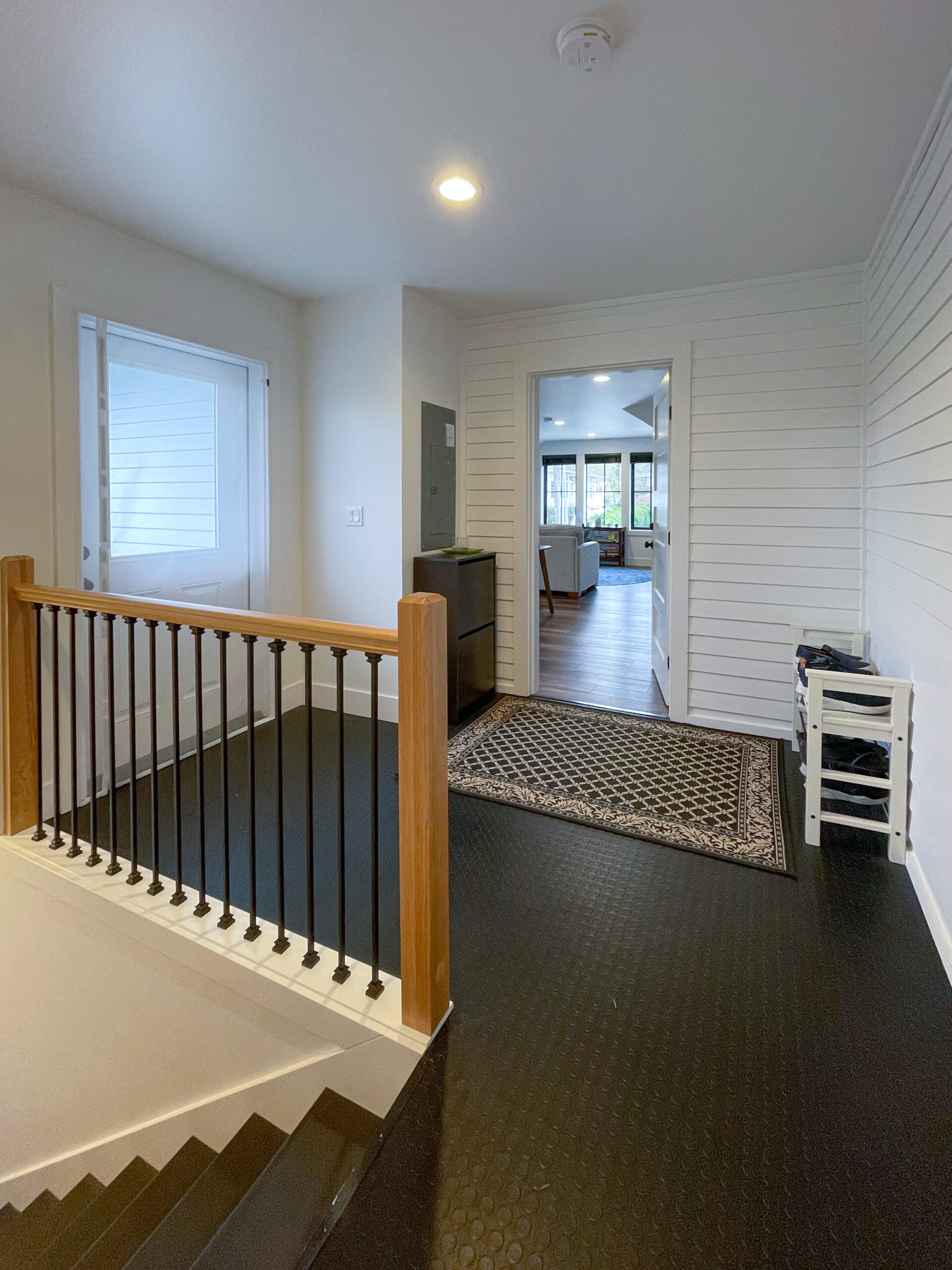 A modern entryway with black flooring, white walls, a wooden stair railing, a small patterned rug, a white shoe rack, and a view into a bright living area with large windows in the background.
