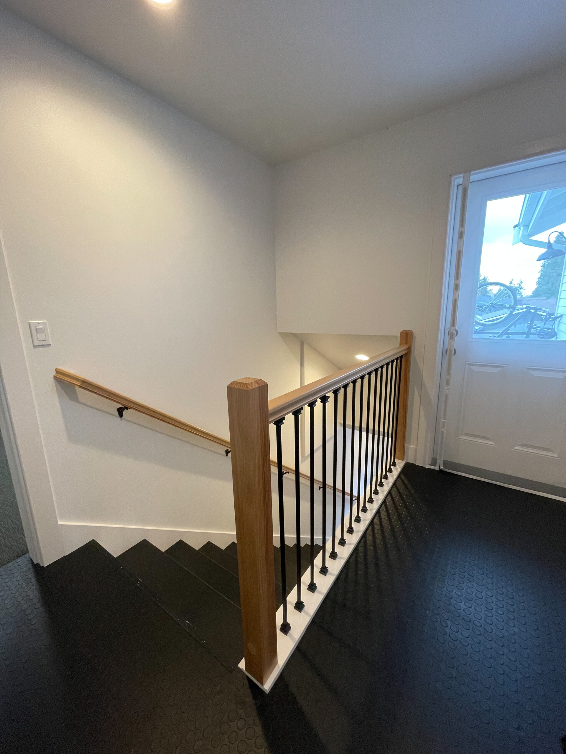A clean, modern stairway with a wooden handrail and black metal balusters leads down from a landing near a white door, with light-colored walls and a dark textured floor.