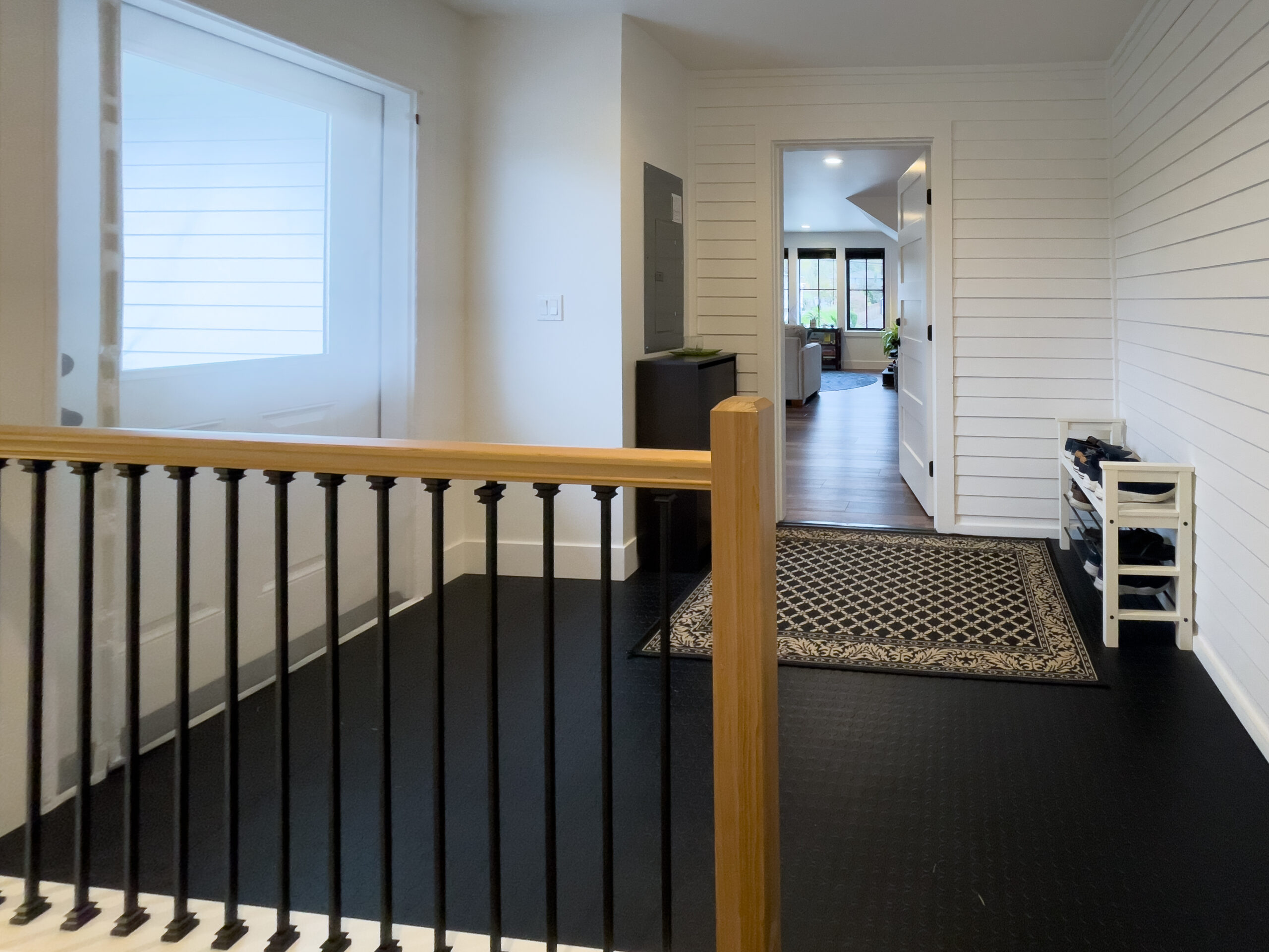 Modern entryway with black floors, white shiplap walls, a wooden handrail with black spindles, a patterned rug, small black cabinet, and a shoe rack; view extends into a bright living area.