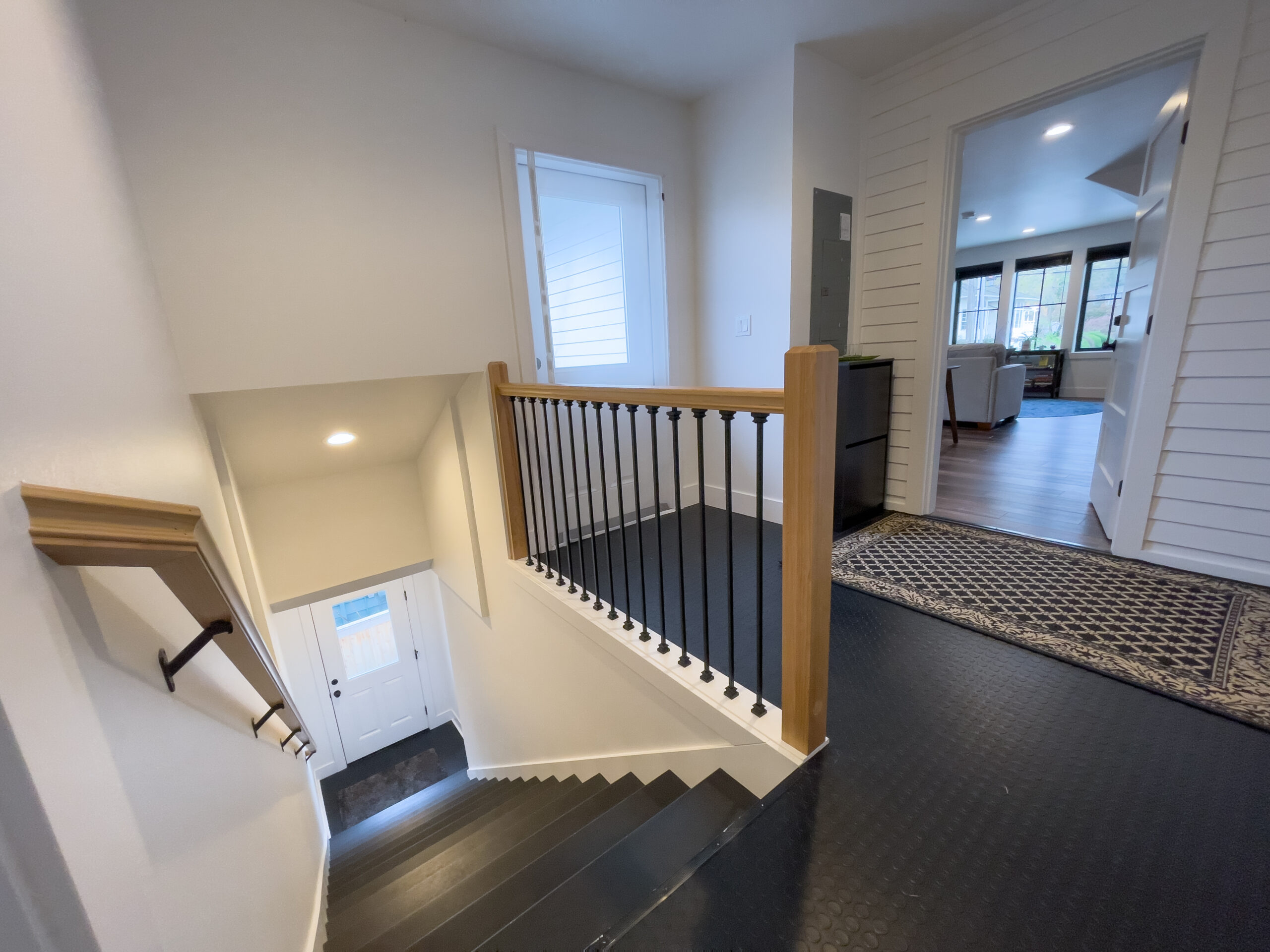 A bright, modern hallway with black metal and wood railing overlooking a stairway leading to a white front door; adjacent rooms with wood floors and large windows are visible.