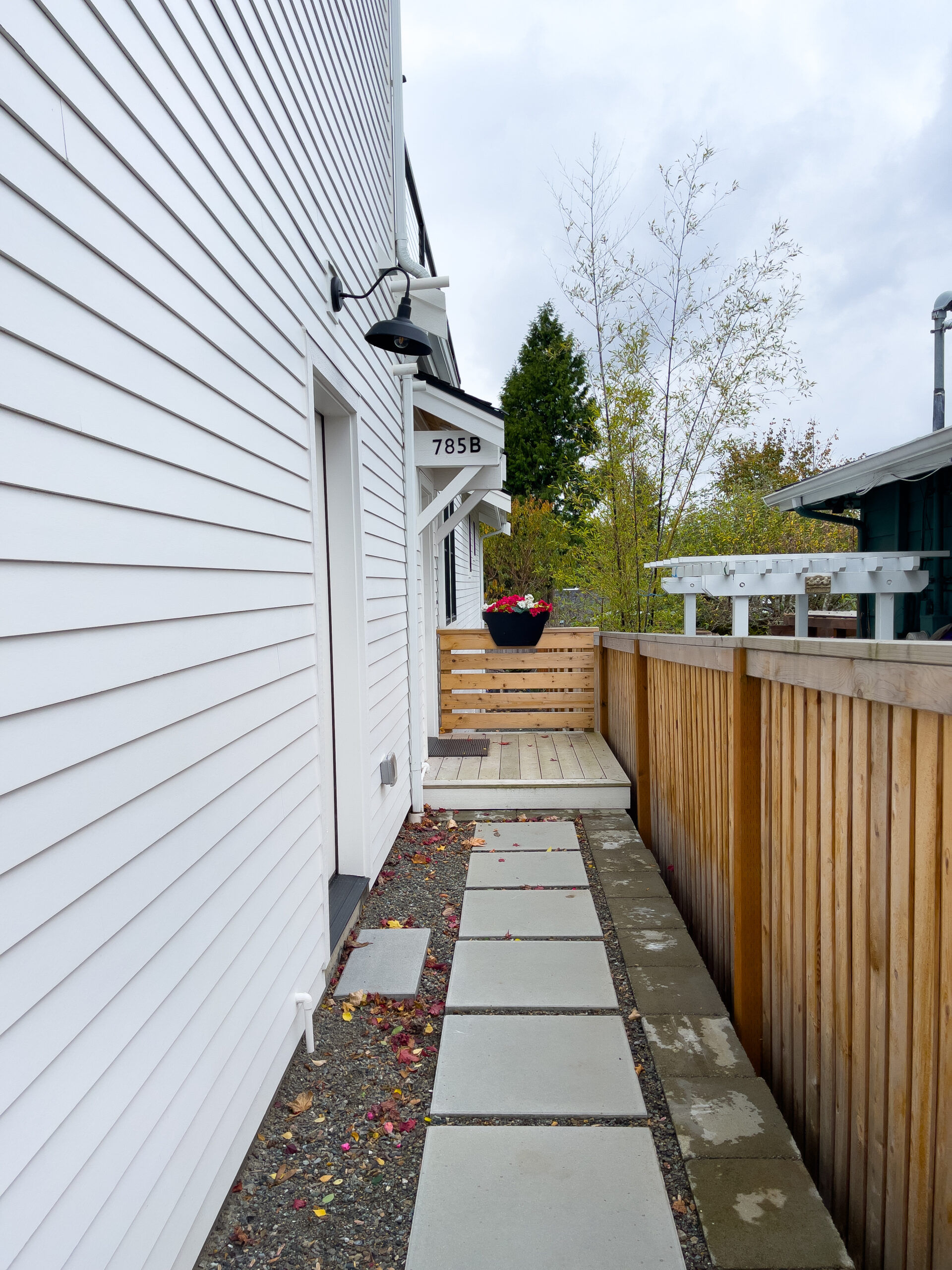 A narrow outdoor walkway with concrete stepping stones runs alongside a white building labeled 785B. A wooden fence borders the right side, and a planter with red flowers sits at the end of the path by a small wooden gate.