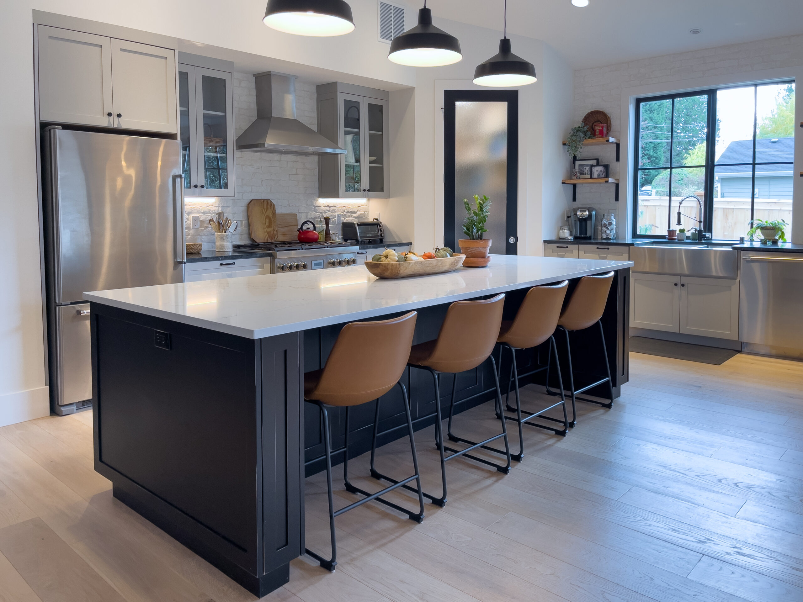 Modern kitchen with a large island featuring brown stools, black base, and a white countertop. Stainless steel appliances, pendant lights, and large windows provide a bright, inviting atmosphere.