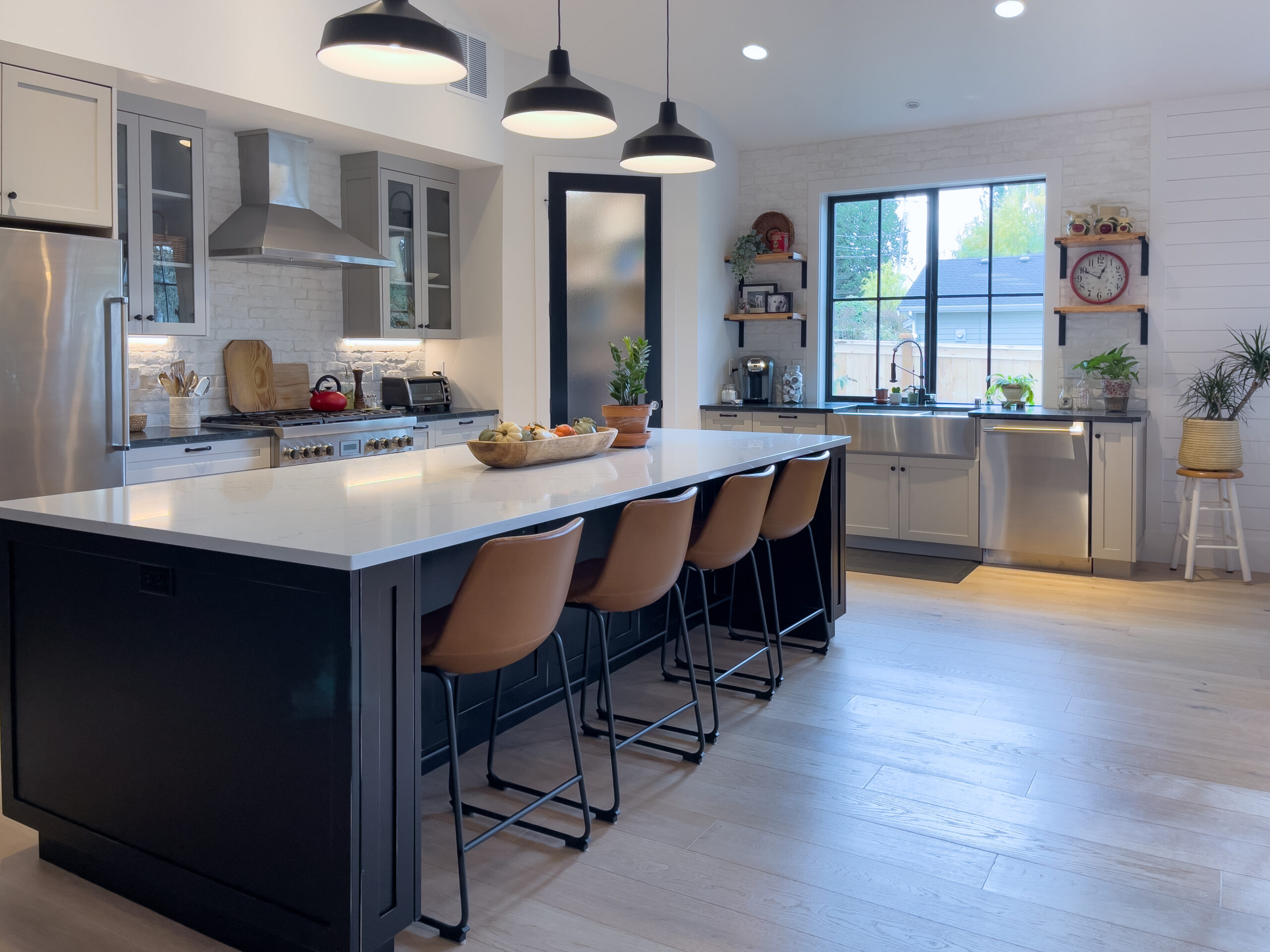 Modern kitchen with a large island, four brown barstools, pendant lights, stainless steel appliances, white cabinets, and a window overlooking a green backyard. Decor includes plants and a red wall clock.
