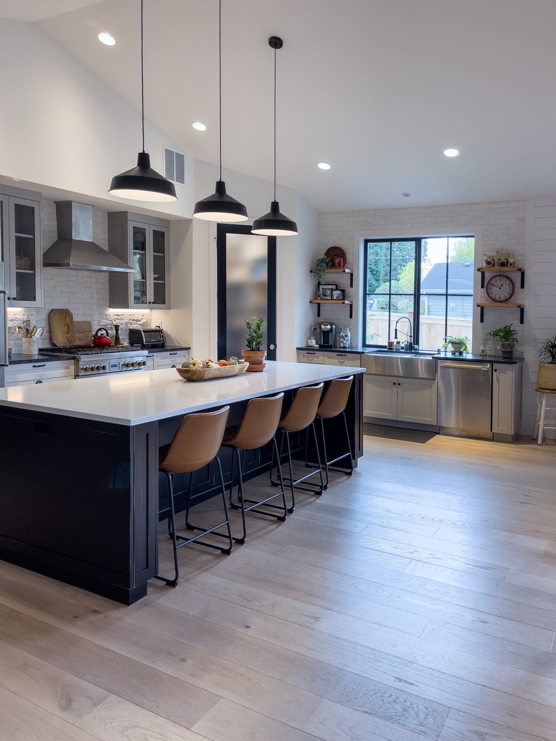 Modern kitchen with a large island, four brown stools, hanging pendant lights, stainless steel appliances, open shelves with plants, and a large window letting in natural light. Light wood flooring throughout.