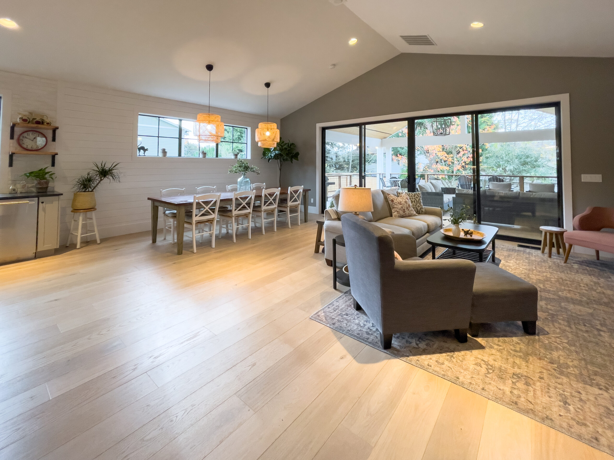 Spacious, modern open-plan dining and living room with light wood floors, large windows, a long dining table with white chairs, cozy seating, and glass doors leading to a patio with greenery outside.