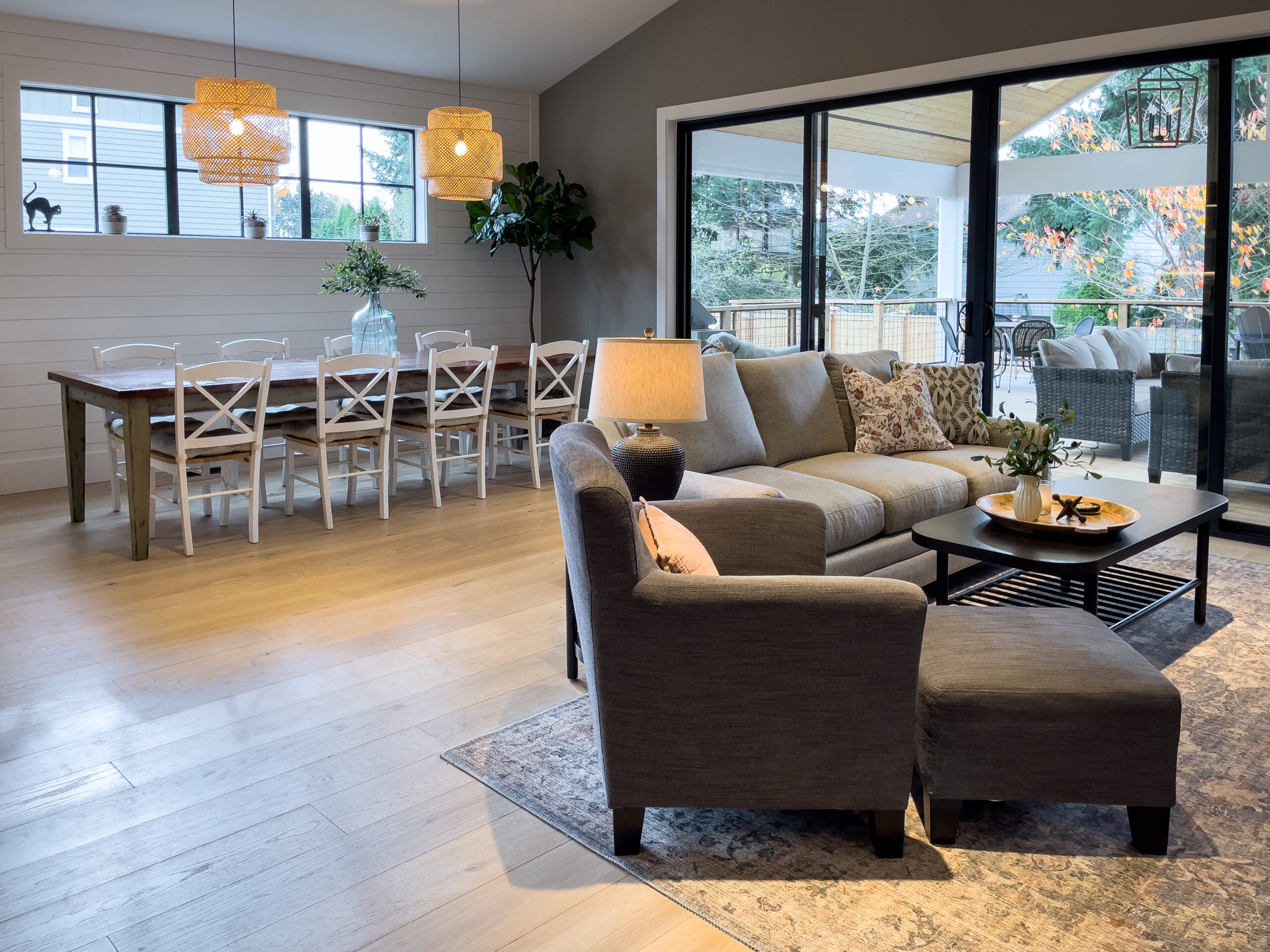 Modern open-concept living and dining area with wood floors, a gray sectional sofa, armchair, rug, and a wooden dining table with white chairs. Large windows and sliding glass doors offer views of trees outside.