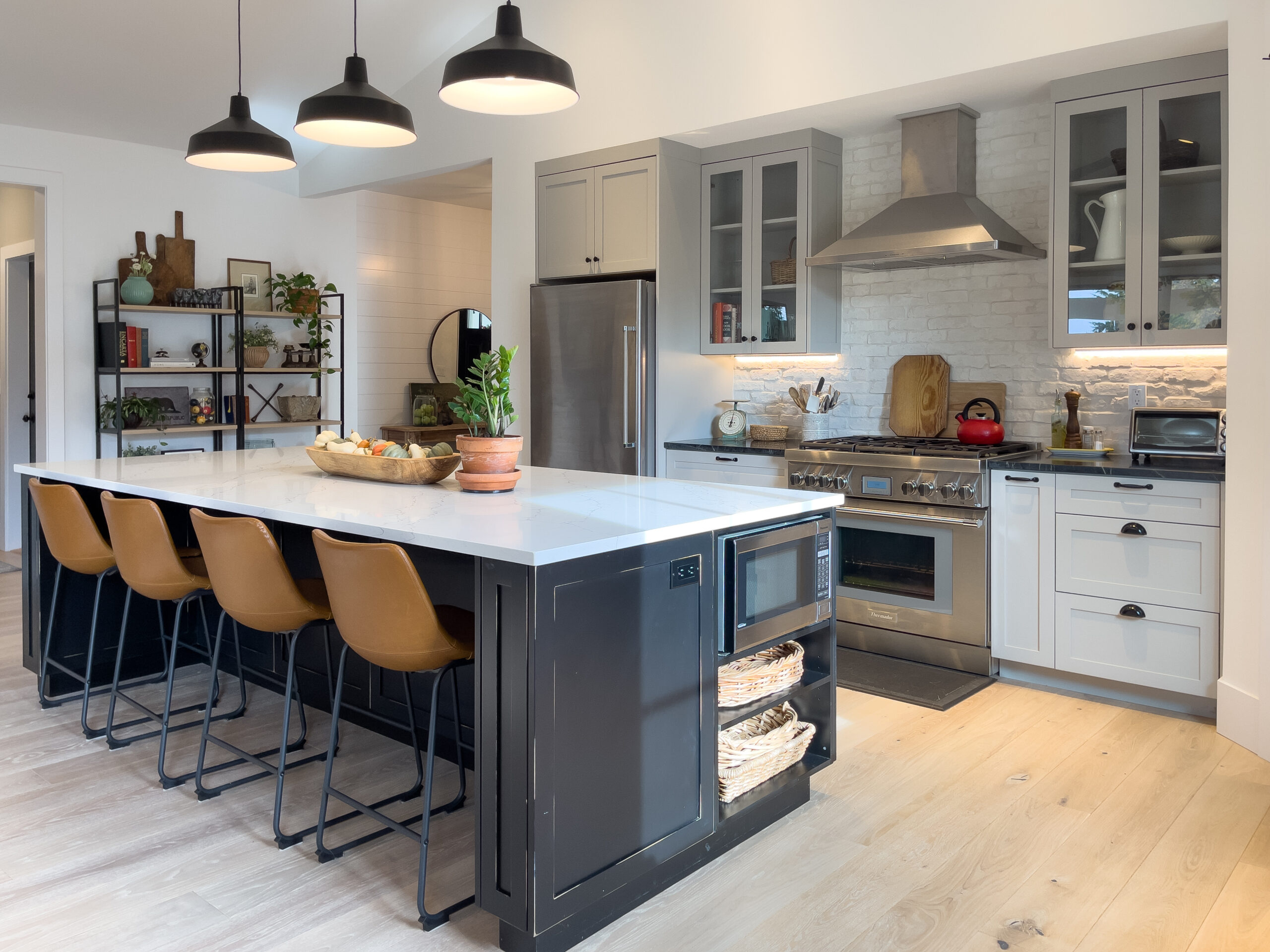 Modern kitchen with a large black island, white countertop, four tan barstools, open shelving with decor, stainless steel appliances, white cabinets, wood flooring, and pendant lights hanging above the island.