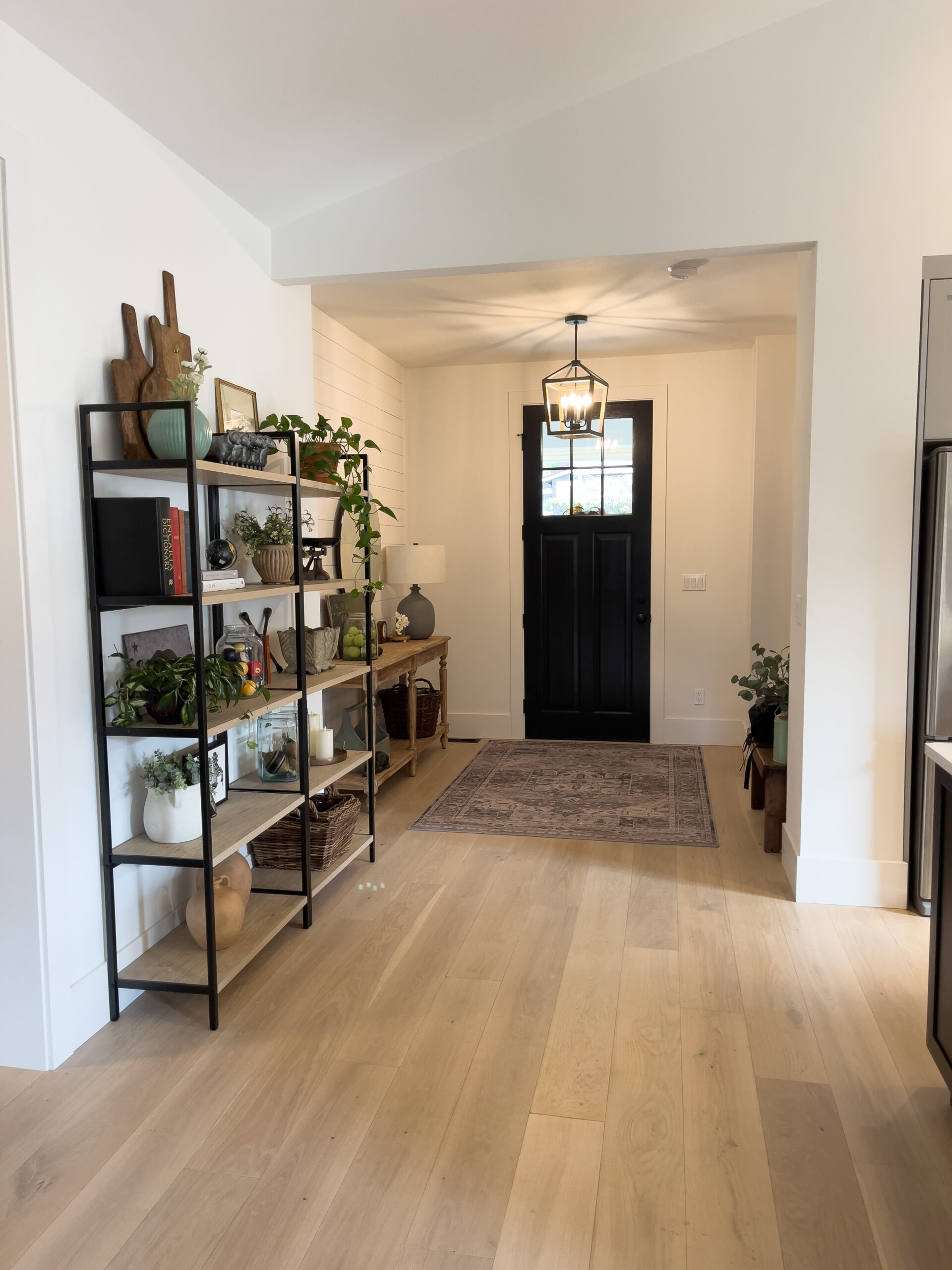 A bright entryway with light wood floors, a black front door, a rug, and shelves on the left holding books, plants, and decor. A table with a lamp and more plants is against the far wall.