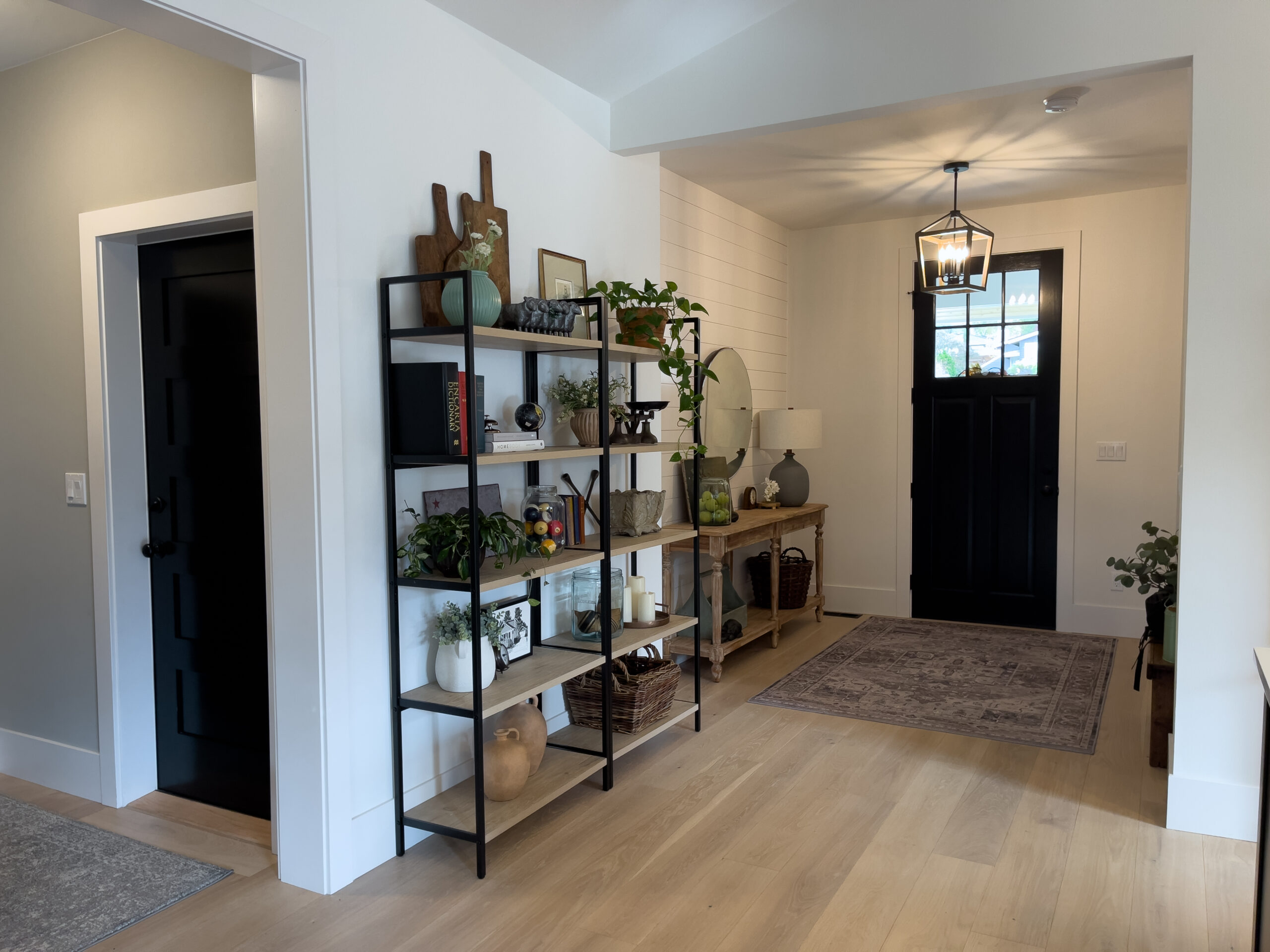 A modern entryway with light wood floors, black doors, a metal and wood bookshelf with plants and decor, a console table with a mirror, and a patterned rug under a geometric light fixture.