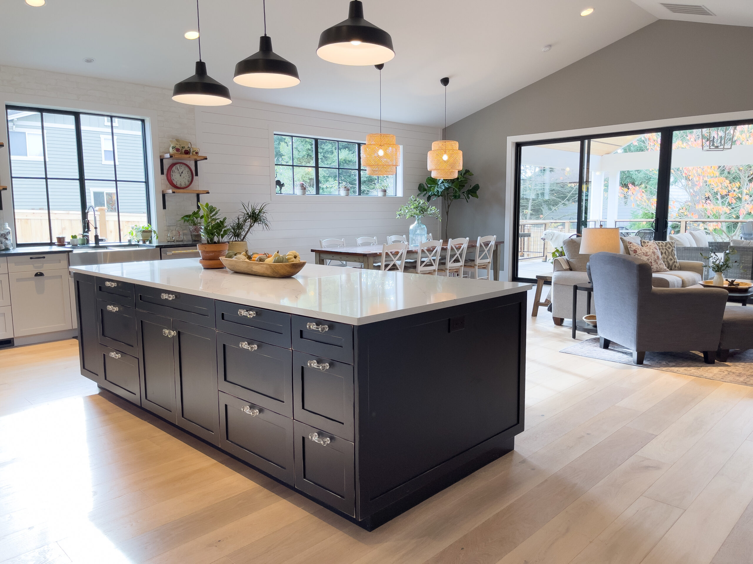 Modern open-concept kitchen and living space with a large black island, white countertops, pendant lights, dining area, and sliding glass doors leading to a patio. Light wood floors and neutral decor create a bright, airy feel.