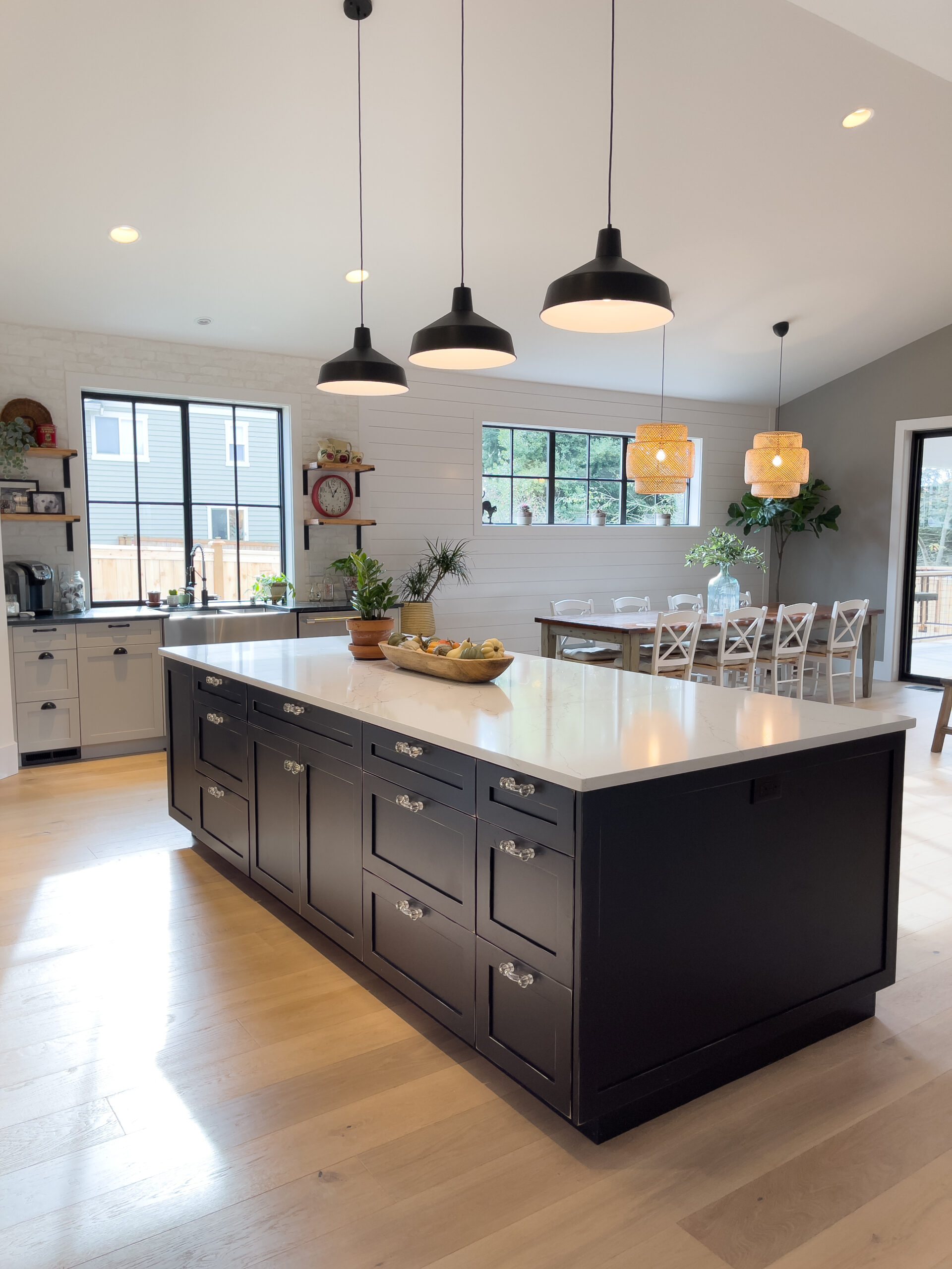 Bright, modern kitchen with a large black island, white countertop, pendant lights, wood floors, and windows letting in natural light. Dining area with a table, chairs, and plants in the background.