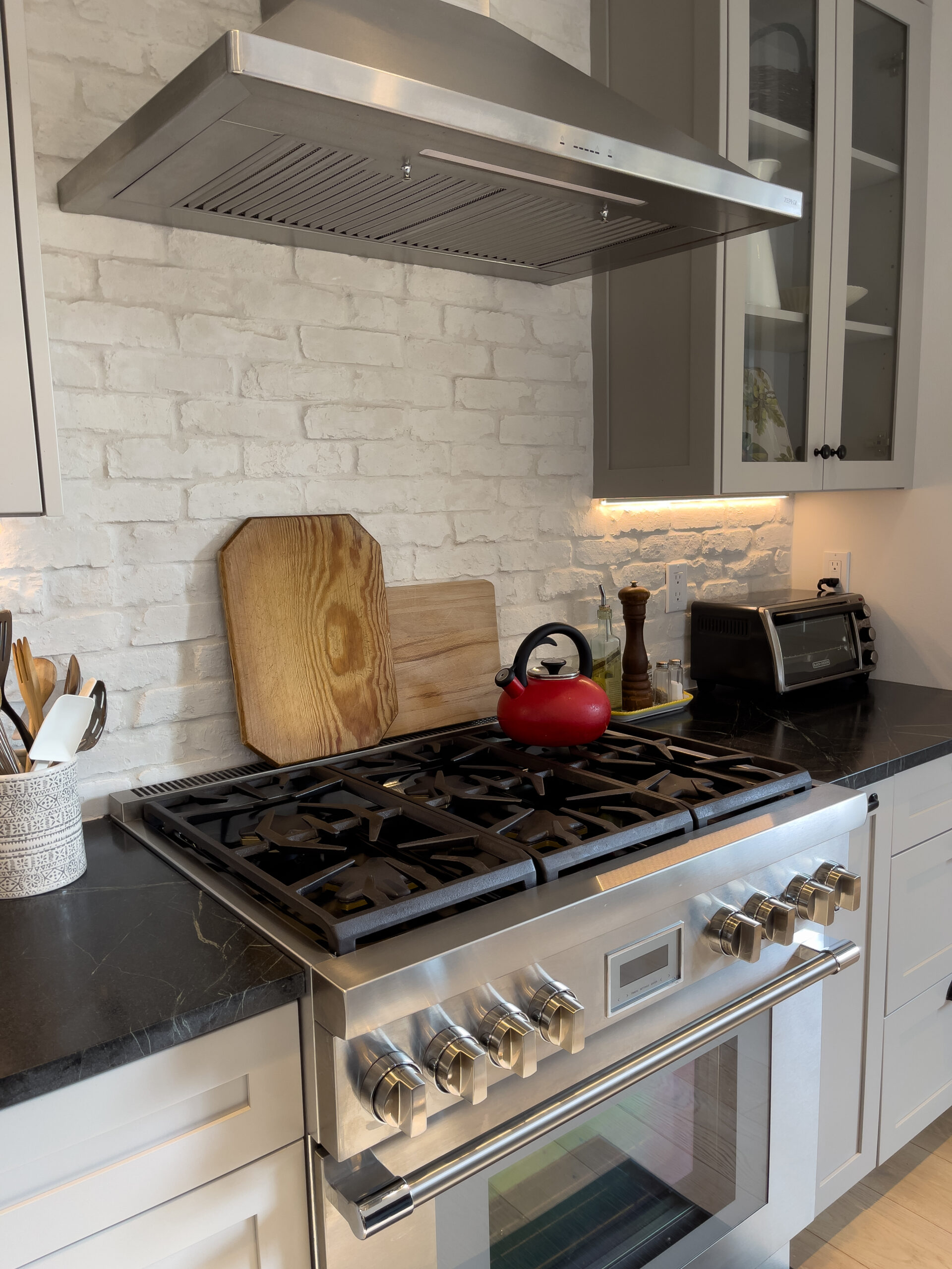 A modern kitchen features a stainless steel gas stove with six burners, a red kettle, wooden cutting boards, utensils in a white holder, a pepper grinder, and a toaster on a dark countertop against a white brick backsplash.