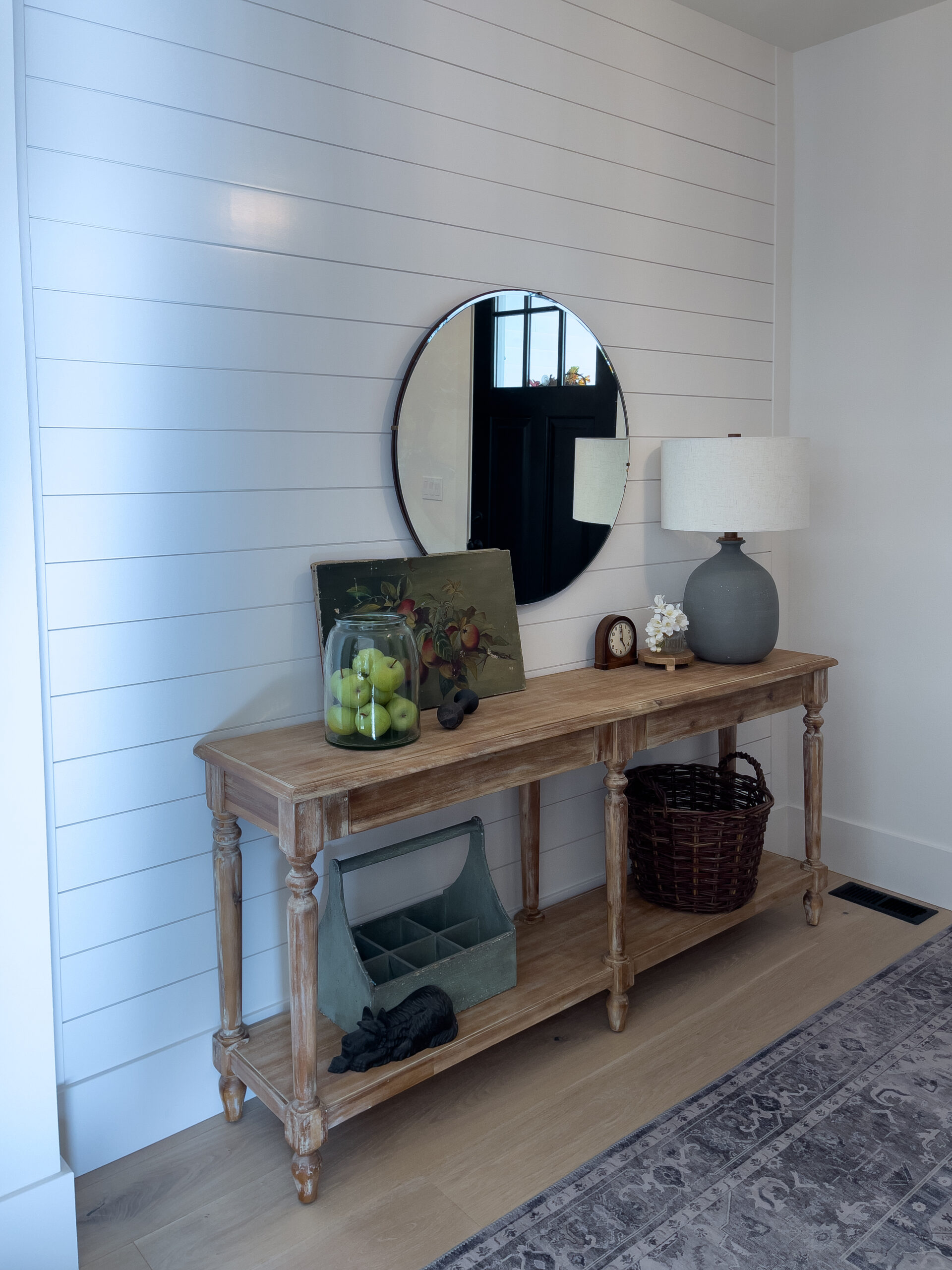 A wooden console table against a white shiplap wall holds a round mirror, lamp, clock, glass jar with green apples, painting, and scattered decor; a basket and storage box sit on the lower shelf.