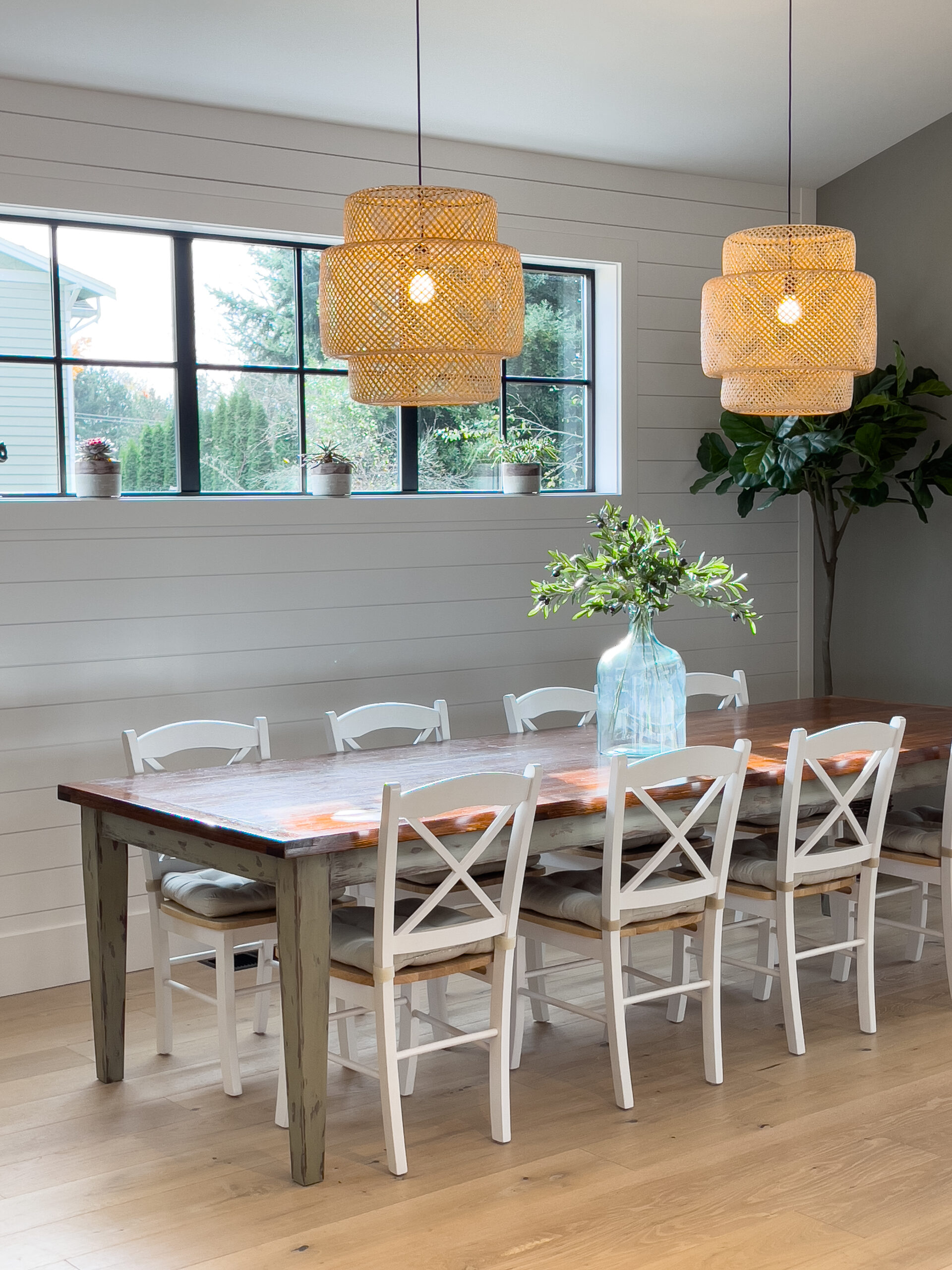Bright dining room with a wooden table, eight white chairs, a clear vase with green branches, two woven pendant lights, large windows, and light wood floors. The space feels modern and inviting.