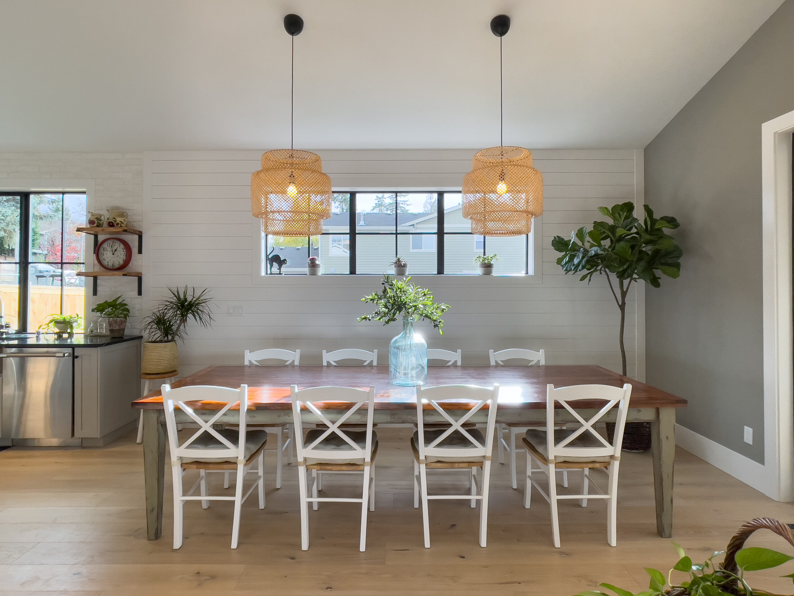 A modern dining room with a long wooden table, eight white chairs, a large glass vase with greenery, two woven pendant lights, plants, and a shiplap accent wall with a window.