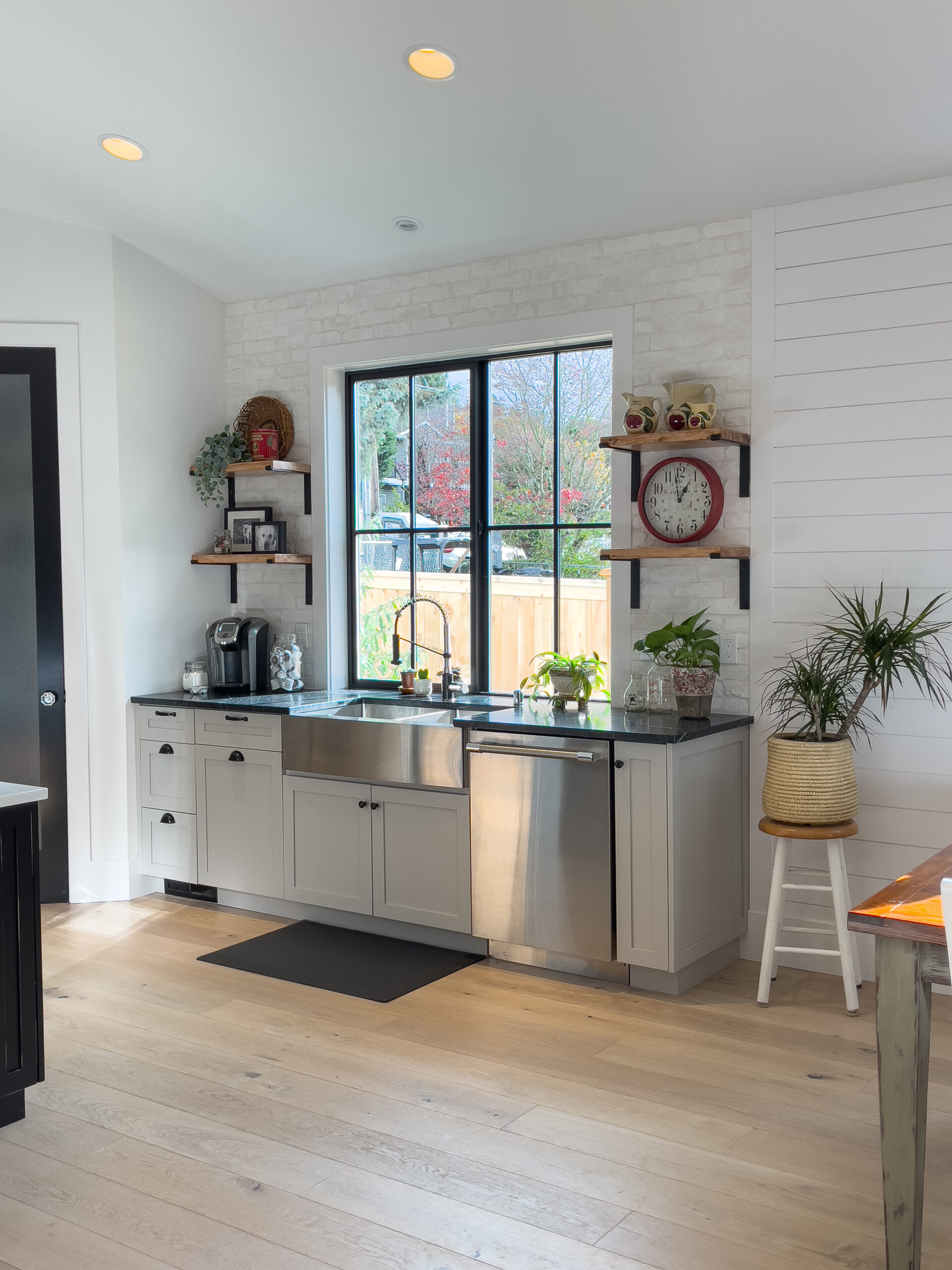 A bright, modern kitchen with light wood floors, white cabinets, a large window above the sink, open shelves with plants and decor, and a wall clock. There is a potted plant on a white stool by the window.
