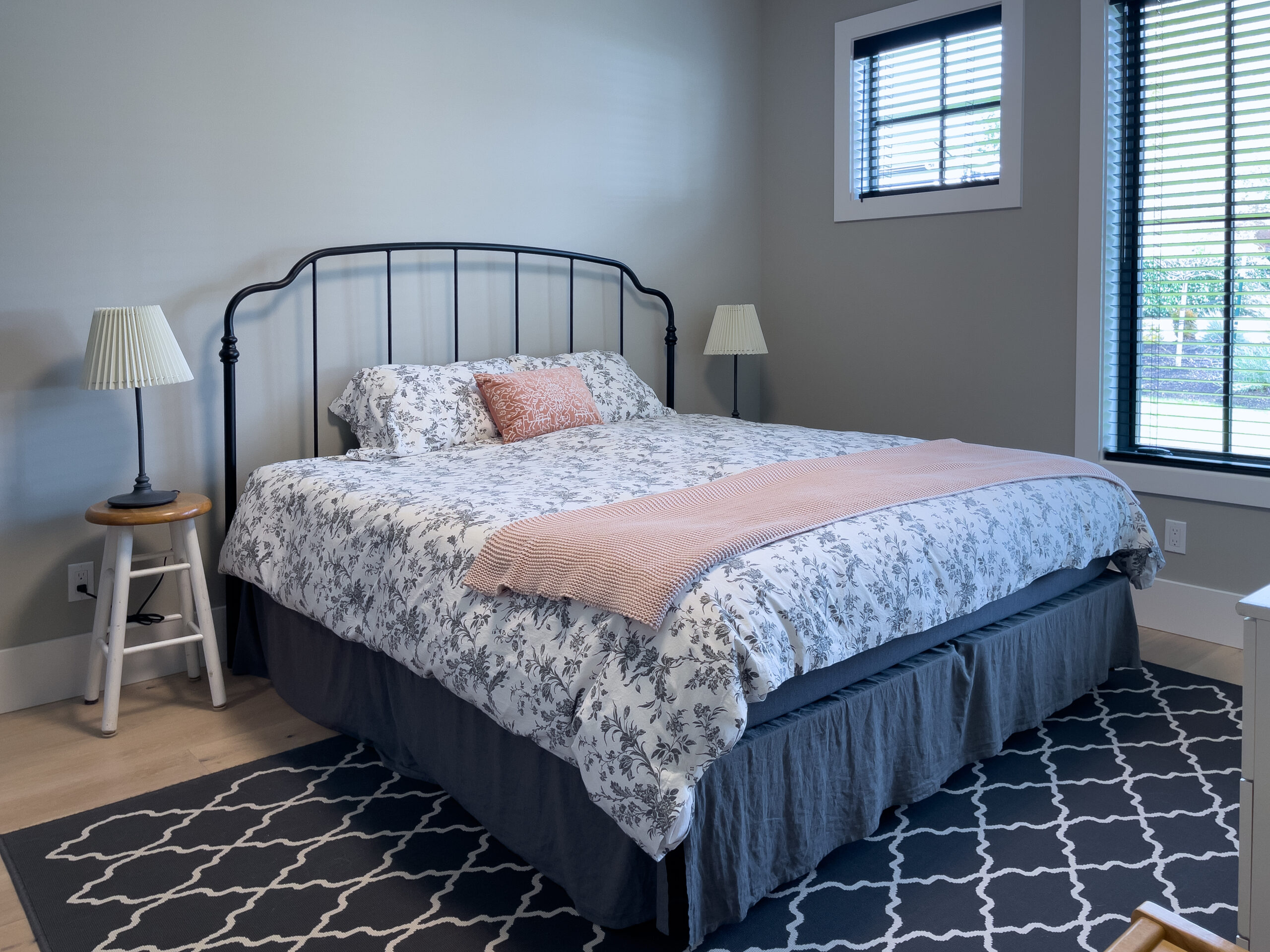 A neatly made bed with floral bedding, two bedside tables with lamps, a soft pink throw blanket, and a patterned rug in a bright bedroom with light-colored walls and two windows.