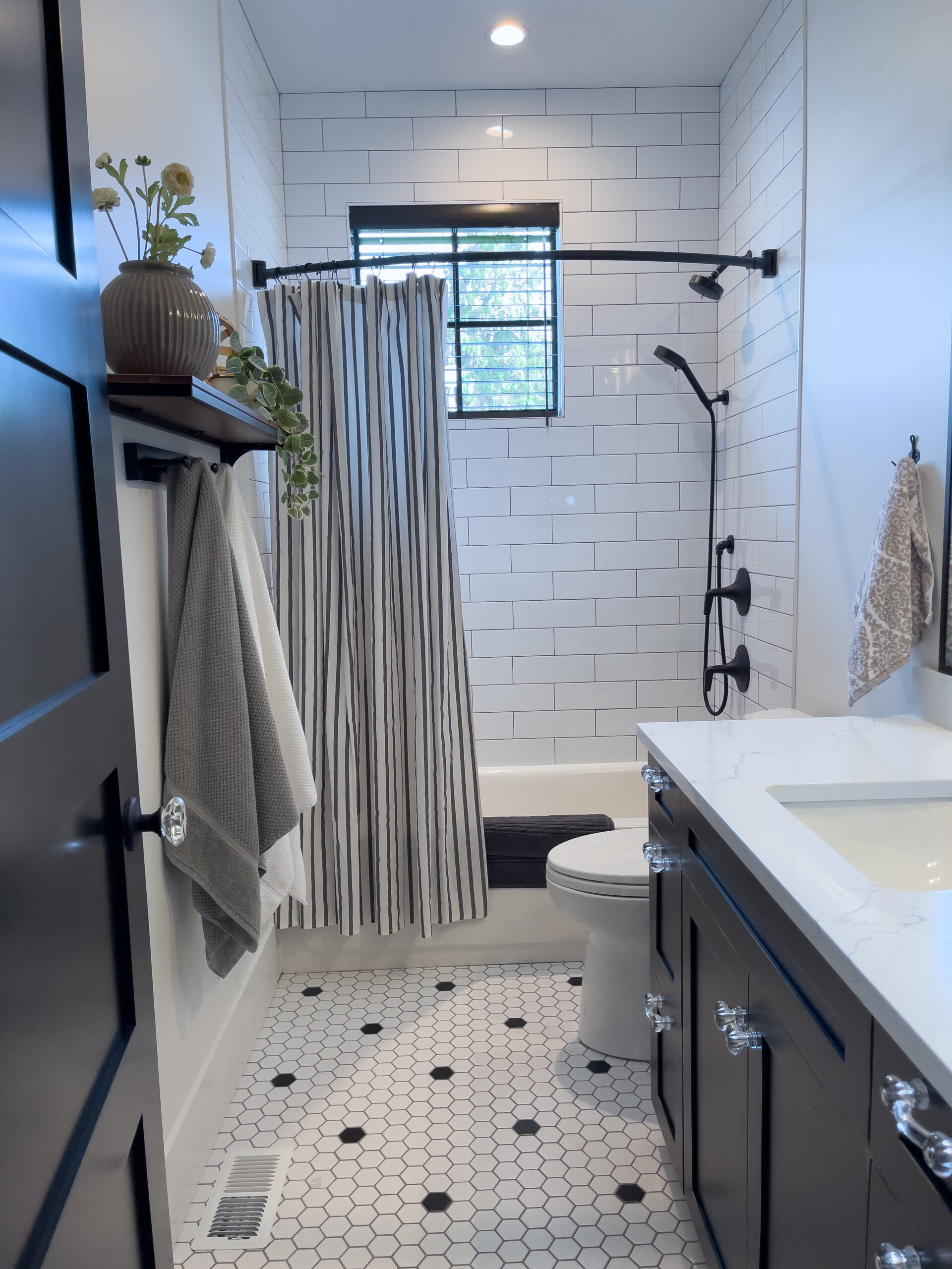 Modern bathroom with white subway tile shower, black fixtures, striped shower curtain, white hexagon floor tiles, dark vanity, white countertop, towel rack with gray towels, and a small plant on a shelf.