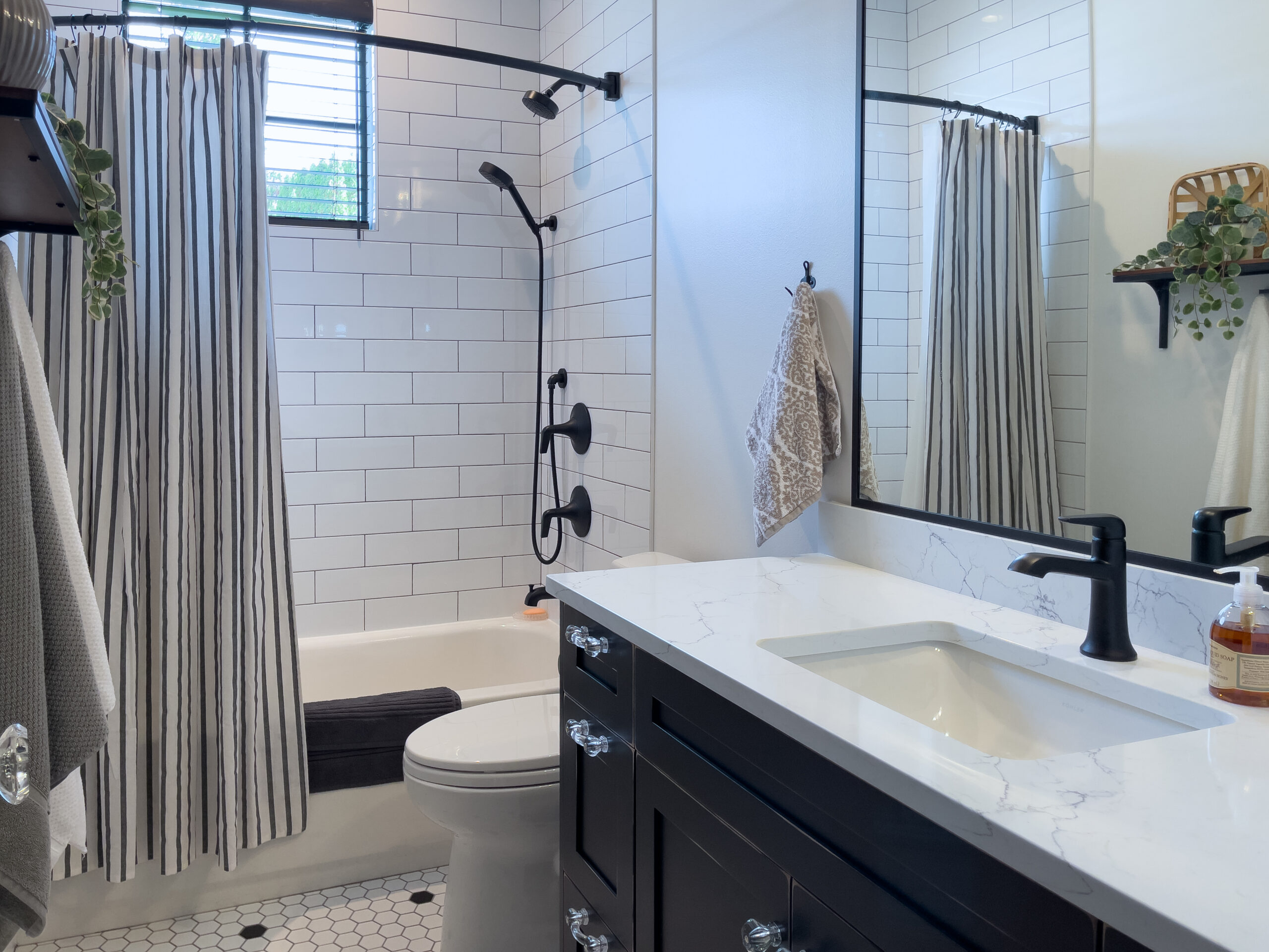 A modern bathroom with white subway tile walls, a black shower fixture, striped shower curtain, black-framed mirror, white countertop with black faucet, hexagonal floor tiles, and a small plant on a shelf.