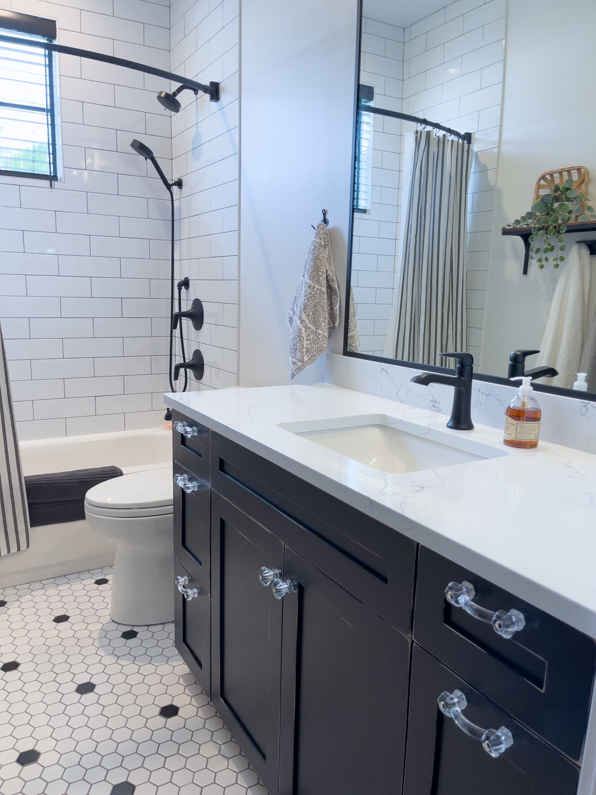 A modern bathroom with white hexagonal tile flooring, a black vanity with a white countertop, a rectangular sink, black fixtures, a bathtub with white subway tiles, a glass shelf, and striped shower curtain.