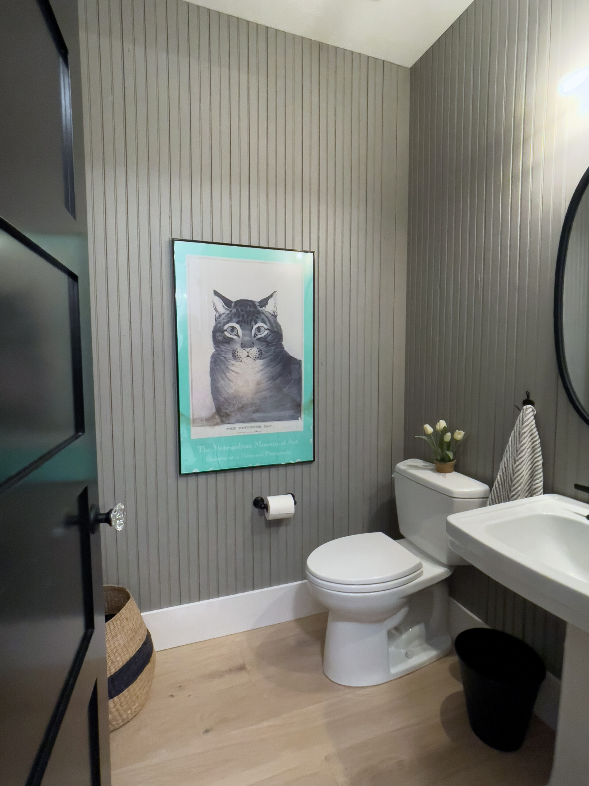 A small bathroom with gray beadboard walls, a white toilet, a white sink, and a framed cat illustration above the toilet. A wicker basket, toilet paper holder, and a striped hand towel are also visible.