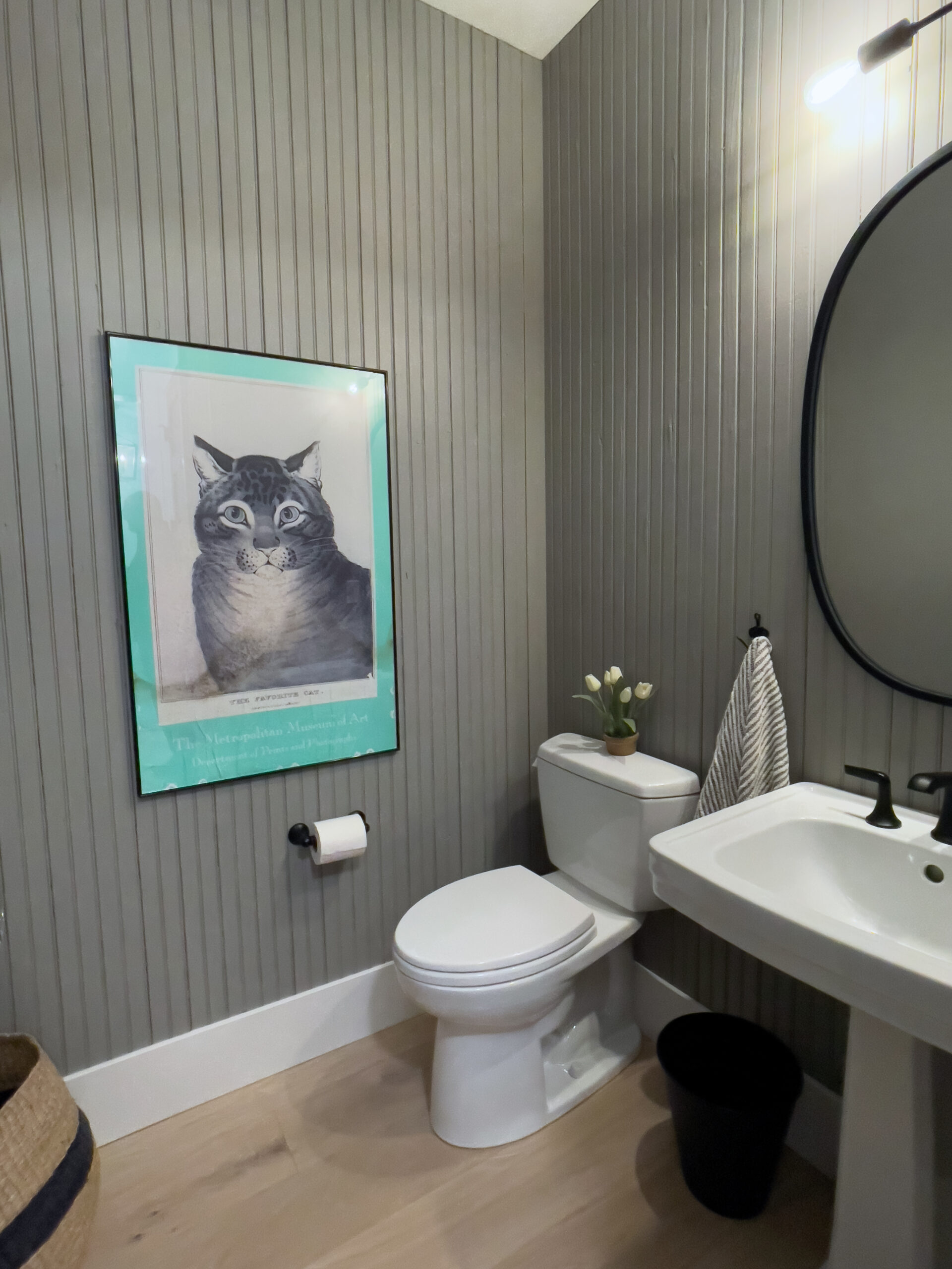 A small bathroom with gray vertical paneling, a white toilet, a white sink, a black oval mirror, a striped towel, a vase of white tulips, and a framed cat artwork on the wall above the toilet.