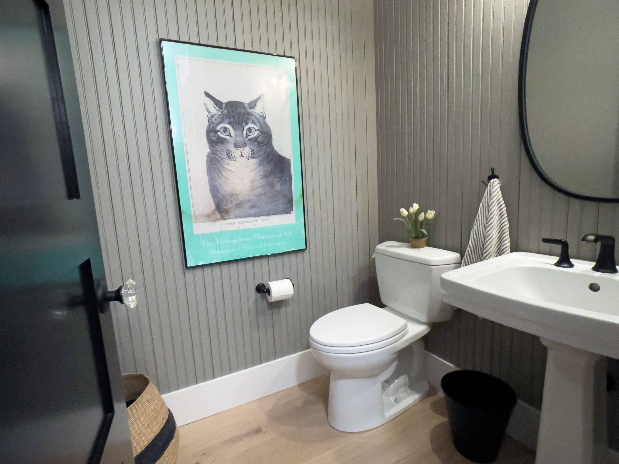 A bathroom with light wood floors, a white toilet, pedestal sink, black fixtures, a small vase with tulips, striped towel, black trash bin, woven basket, and a framed cat illustration on the wall.