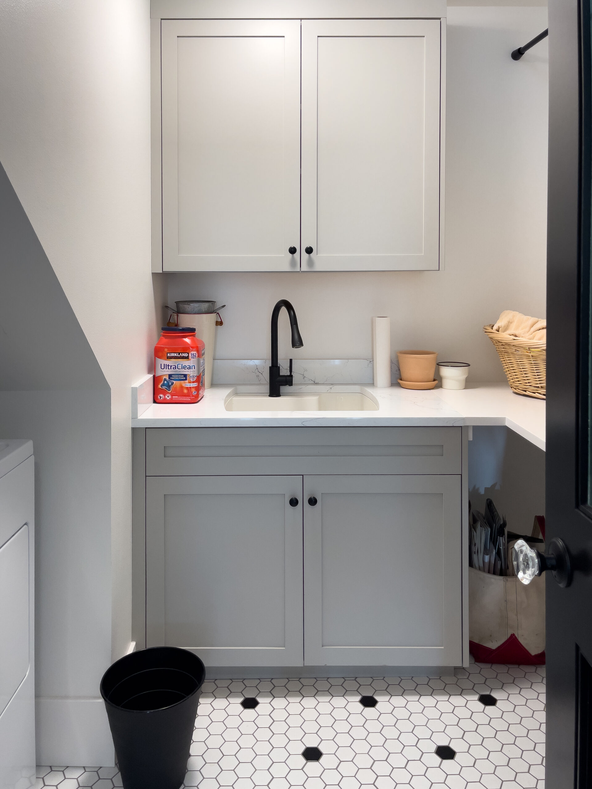 A laundry room with white cabinets, a sink, black faucet, detergent, paper towels, a black trash bin, laundry basket, and hexagonal white tile floor. The washer is partially visible on the left.