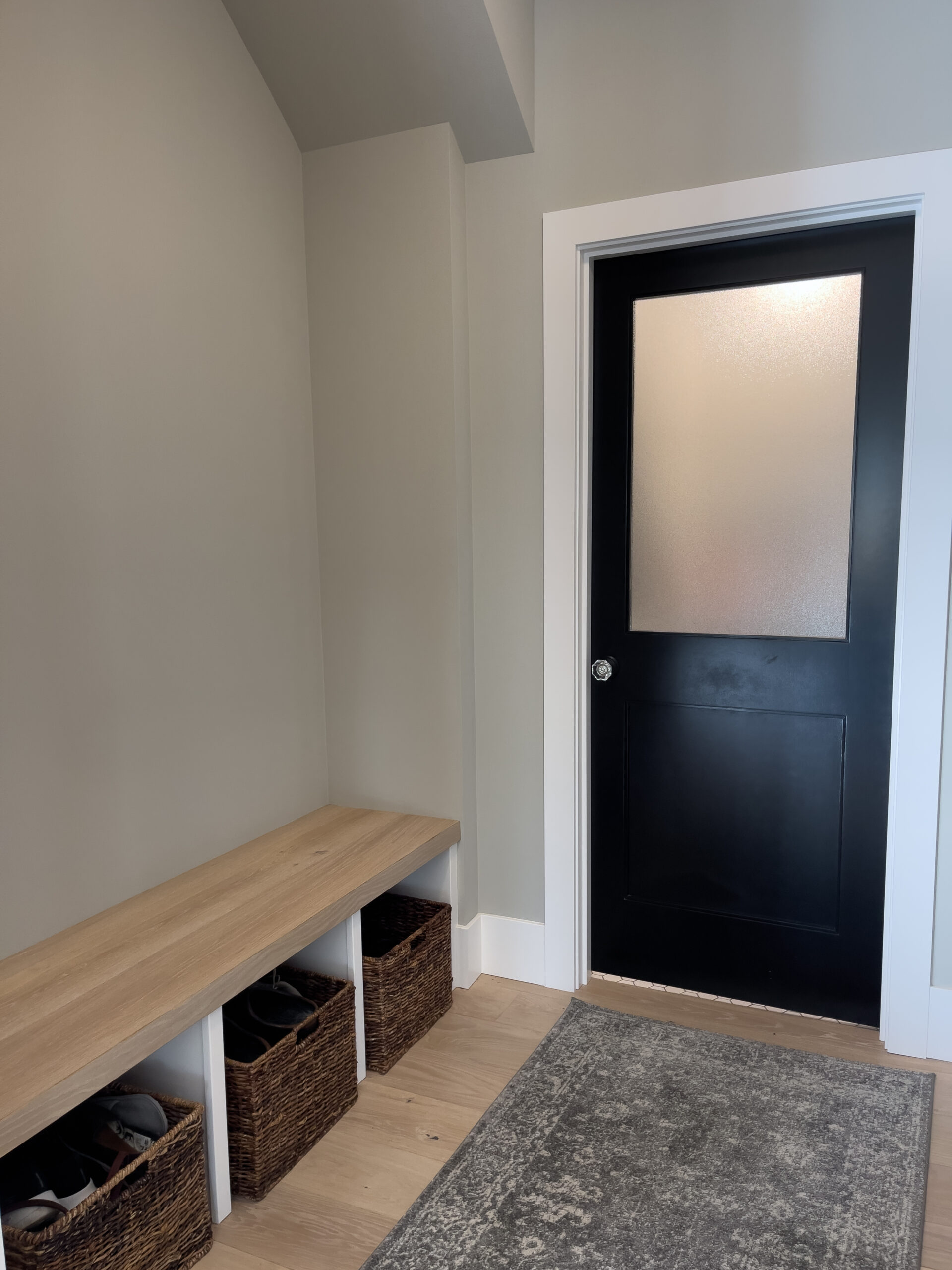 A modern entryway with light gray walls, a black door with frosted glass, a wooden bench with cubbies holding wicker baskets, and a gray rug on a light wood floor.