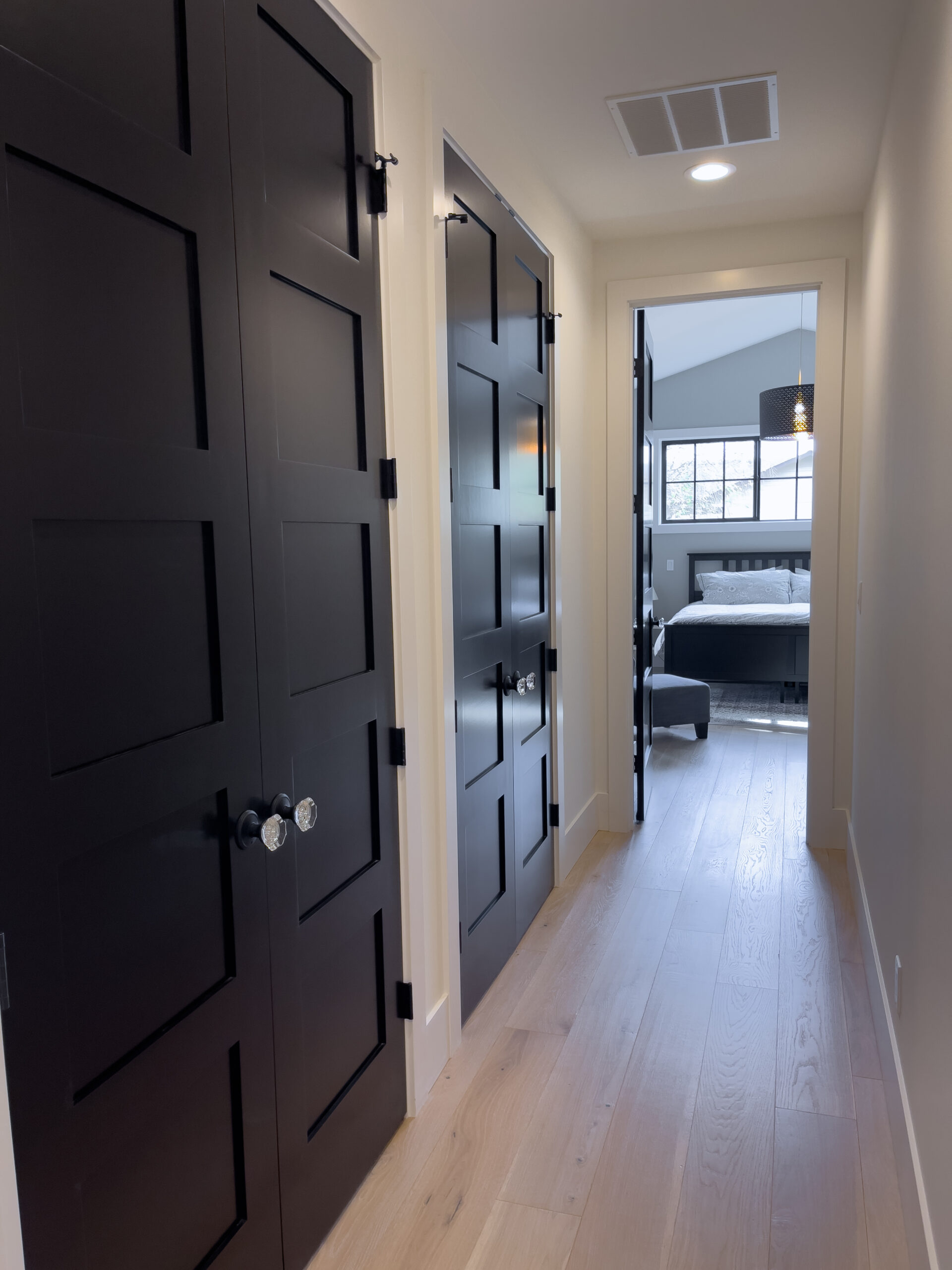 A bright hallway with light wood floors, featuring two black paneled doors on the left and leading to a bedroom with a large bed and windows letting in natural light.
