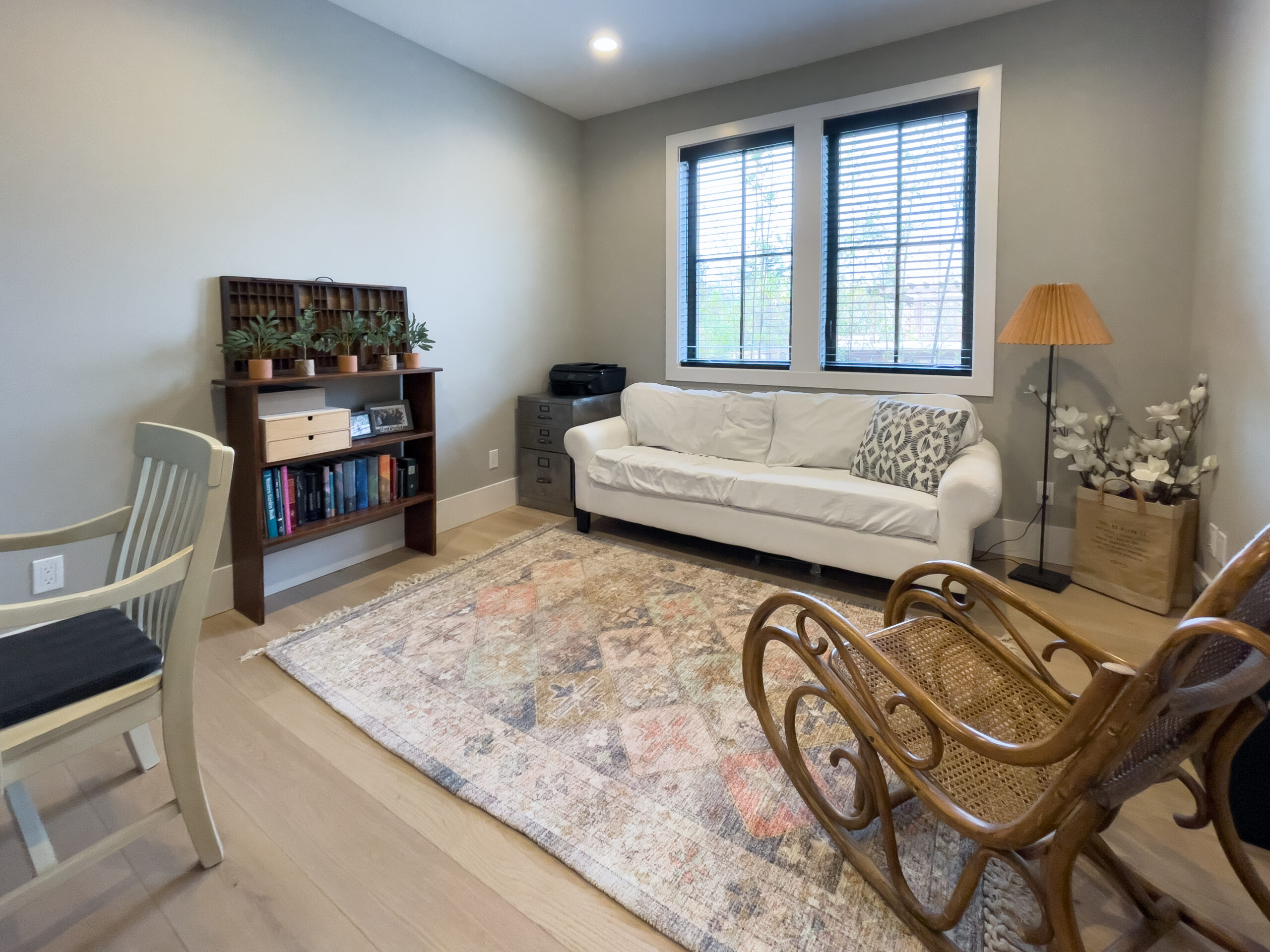 A cozy living room with a white sofa, patterned pillows, a wooden rocking chair, a beige rug, a bookshelf with plants and books, a lamp, and two windows with blinds letting in natural light.