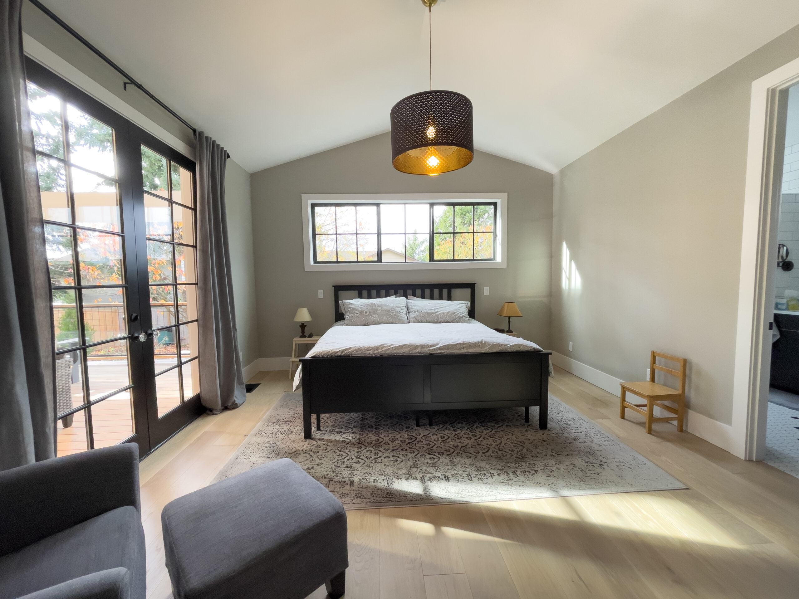 A bright bedroom with a large black bed, beige walls, a patterned rug, a black pendant light, two bedside tables with lamps, a gray armchair, and floor-to-ceiling windows opening to a deck.