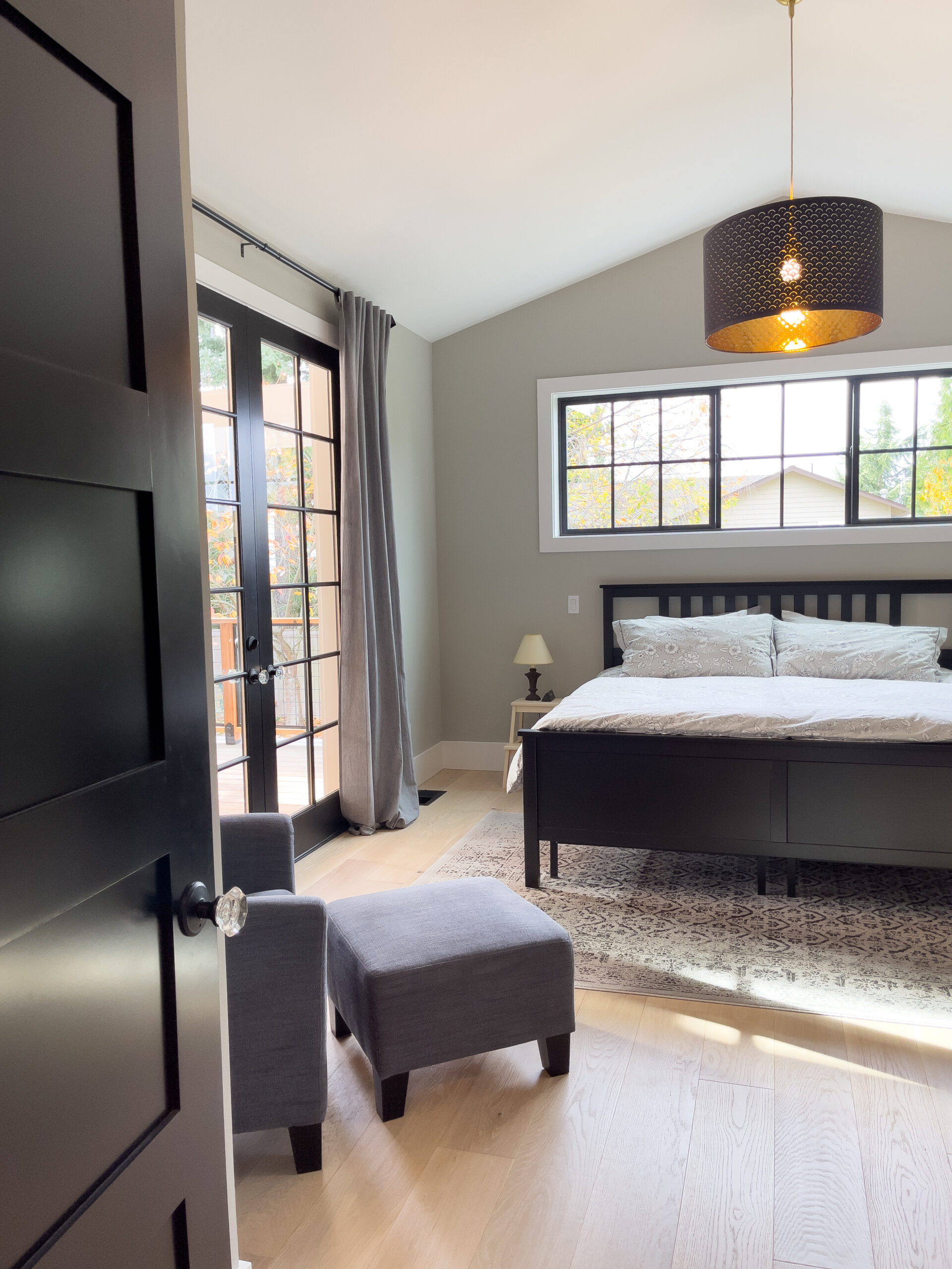 A bright, modern bedroom with a large bed, gray walls, wood floors, a black pendant light, a patterned rug, and a gray armchair with ottoman near glass doors and tall windows.