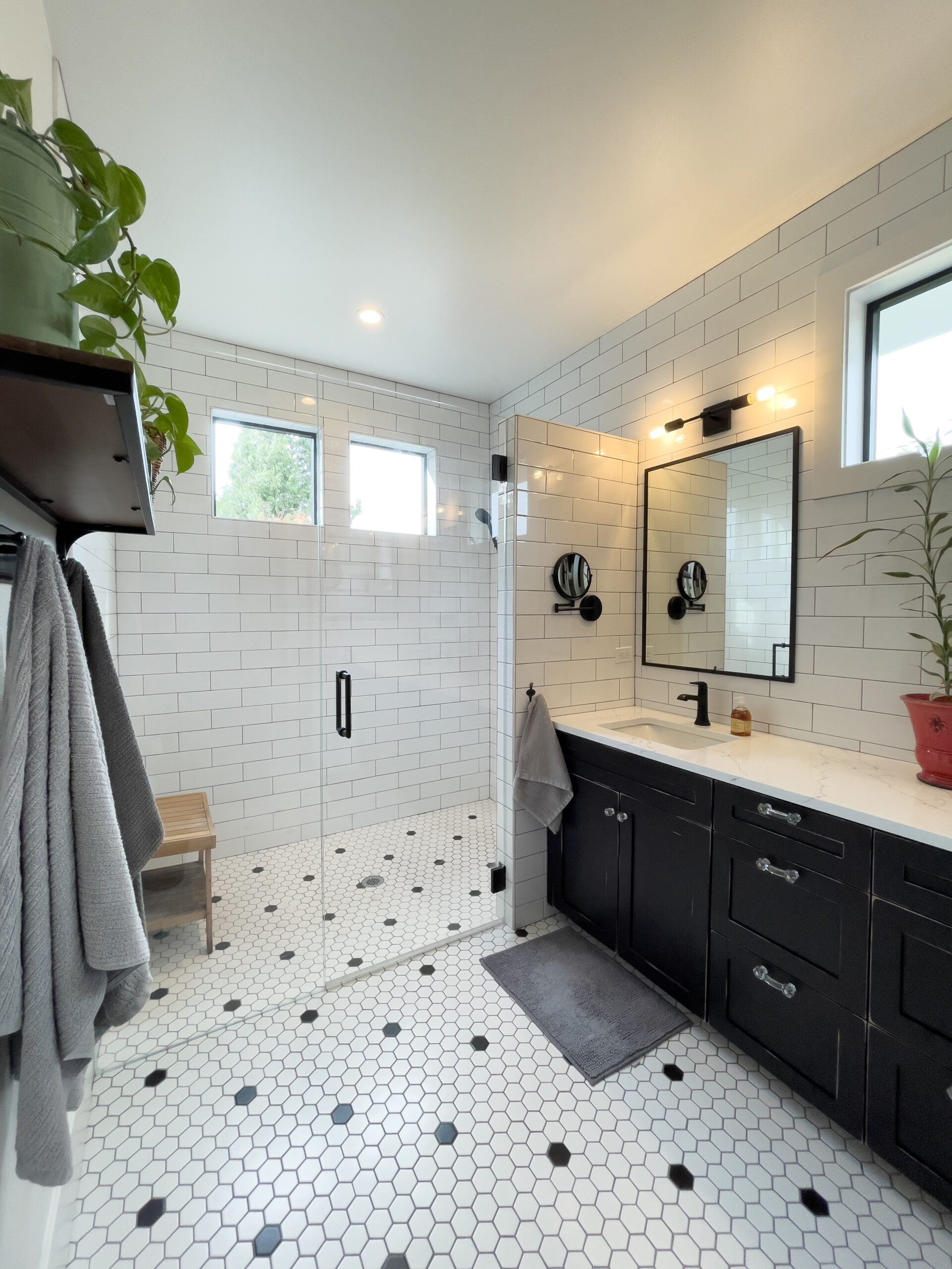 Modern bathroom with white subway tile walls, black vanity, hexagonal white floor tiles with black accents, glass shower enclosure, wall-mounted mirror, towels, and potted plants for decoration.