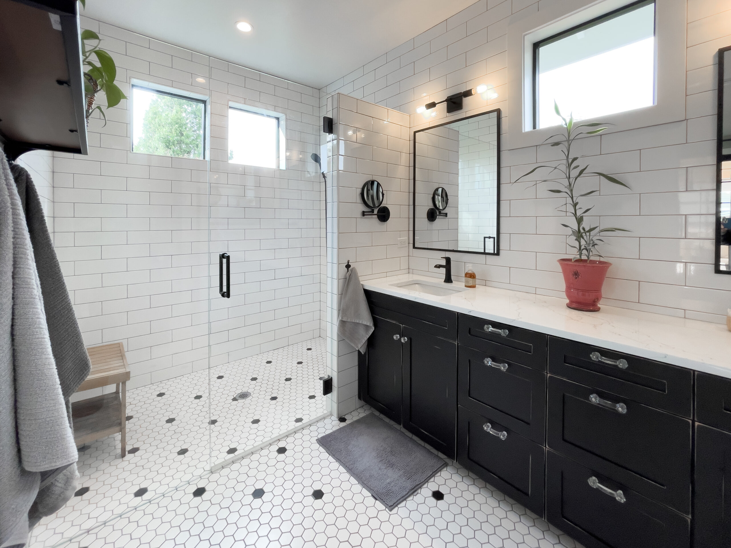 Modern bathroom with white subway tile walls, hexagonal white tile floor, a glass shower, black vanity with double sinks, rectangular mirrors, a potted plant, and towels hanging on hooks.