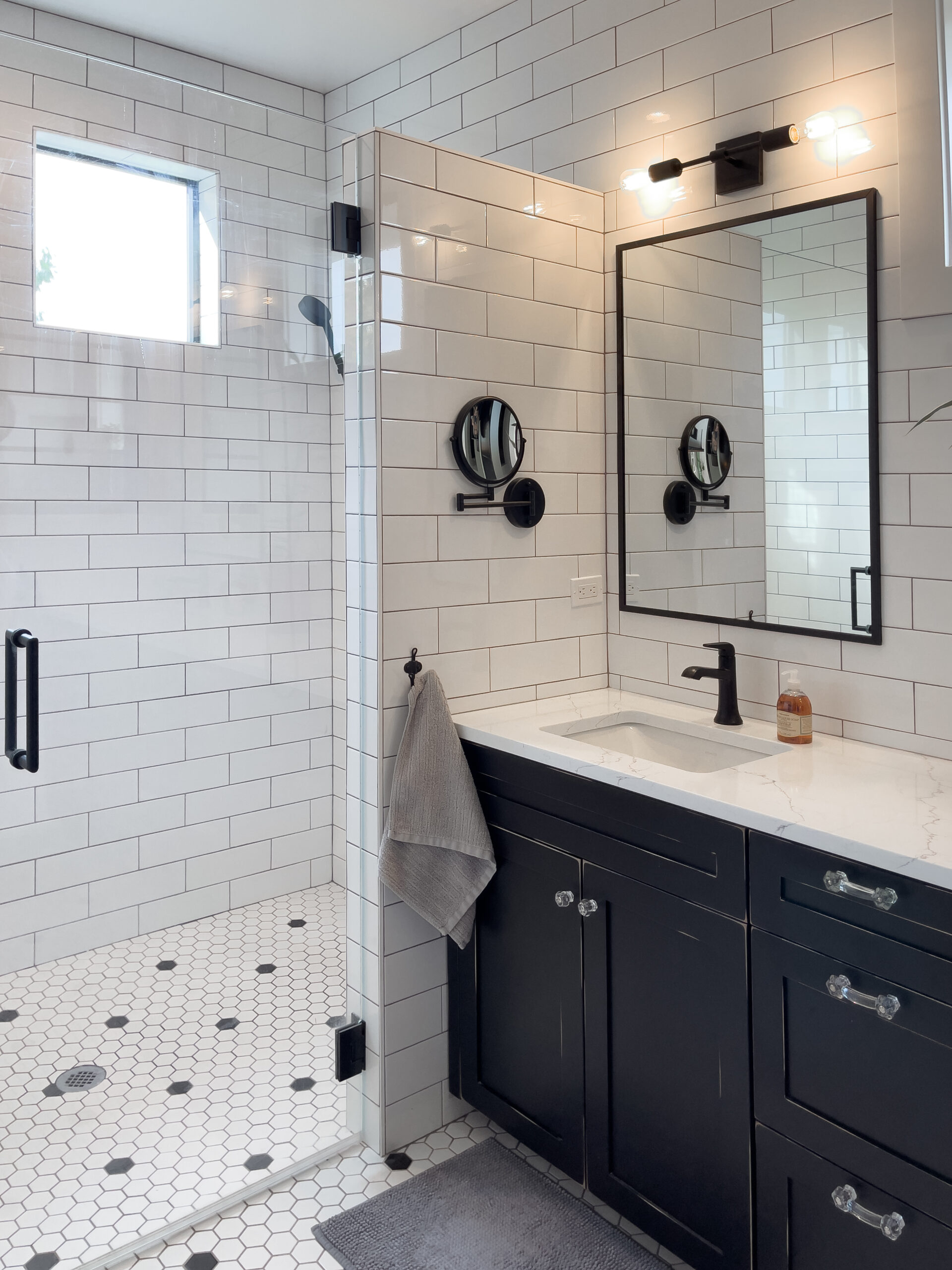 Modern bathroom with white subway tile walls, a glass-enclosed shower with hexagonal floor tiles, a black-framed mirror, a black vanity with white countertop, wall-mounted faucets, and a gray towel hanging by the sink.