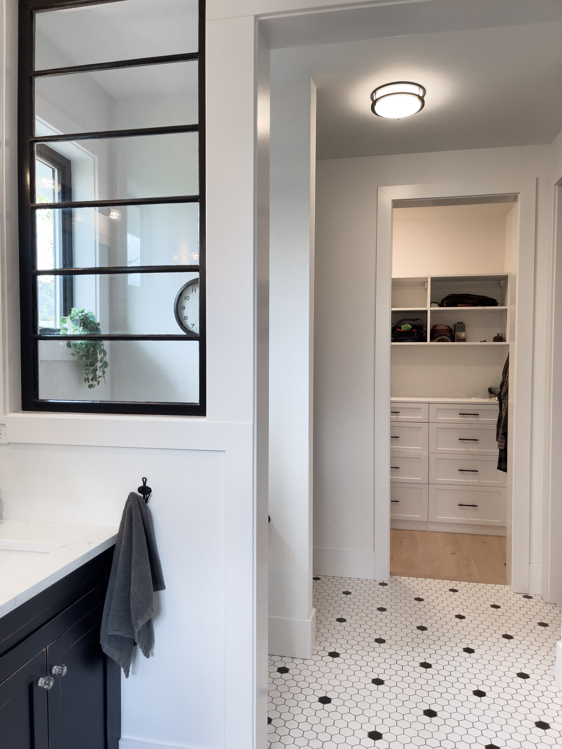 Modern bathroom with white walls and black trim, featuring a towel hanging on a black hook, a window with black grid frame, hexagon tile flooring, and a walk-in closet with shelves and drawers in the background.